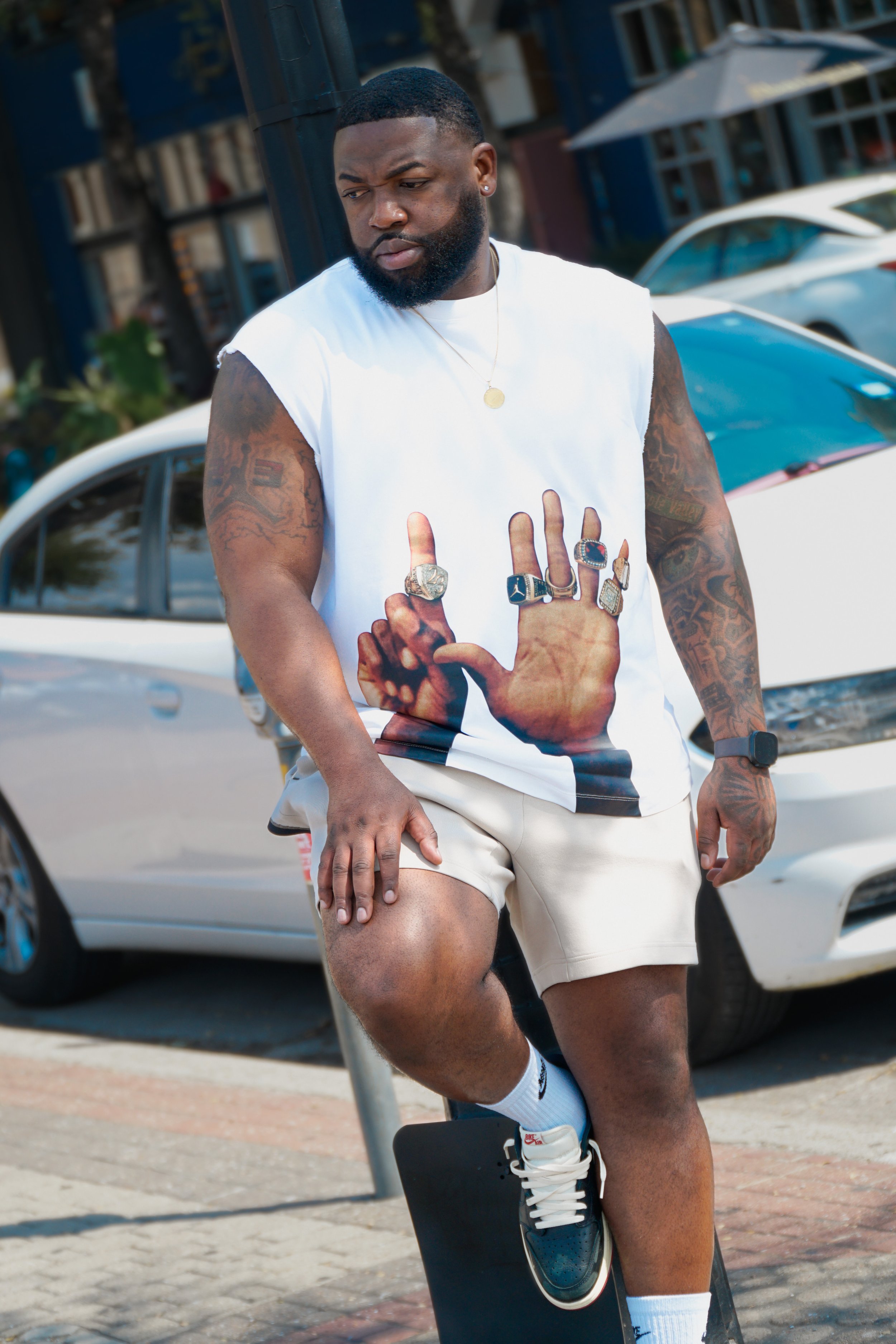 A man with tattoos on his arms and beard wearing a sleeveless T-shirt with a print of hands displaying rings. He has rings on his fingers, a necklace, a watch, and is wearing sneakers and shorts. He is standing outdoors near parked cars.