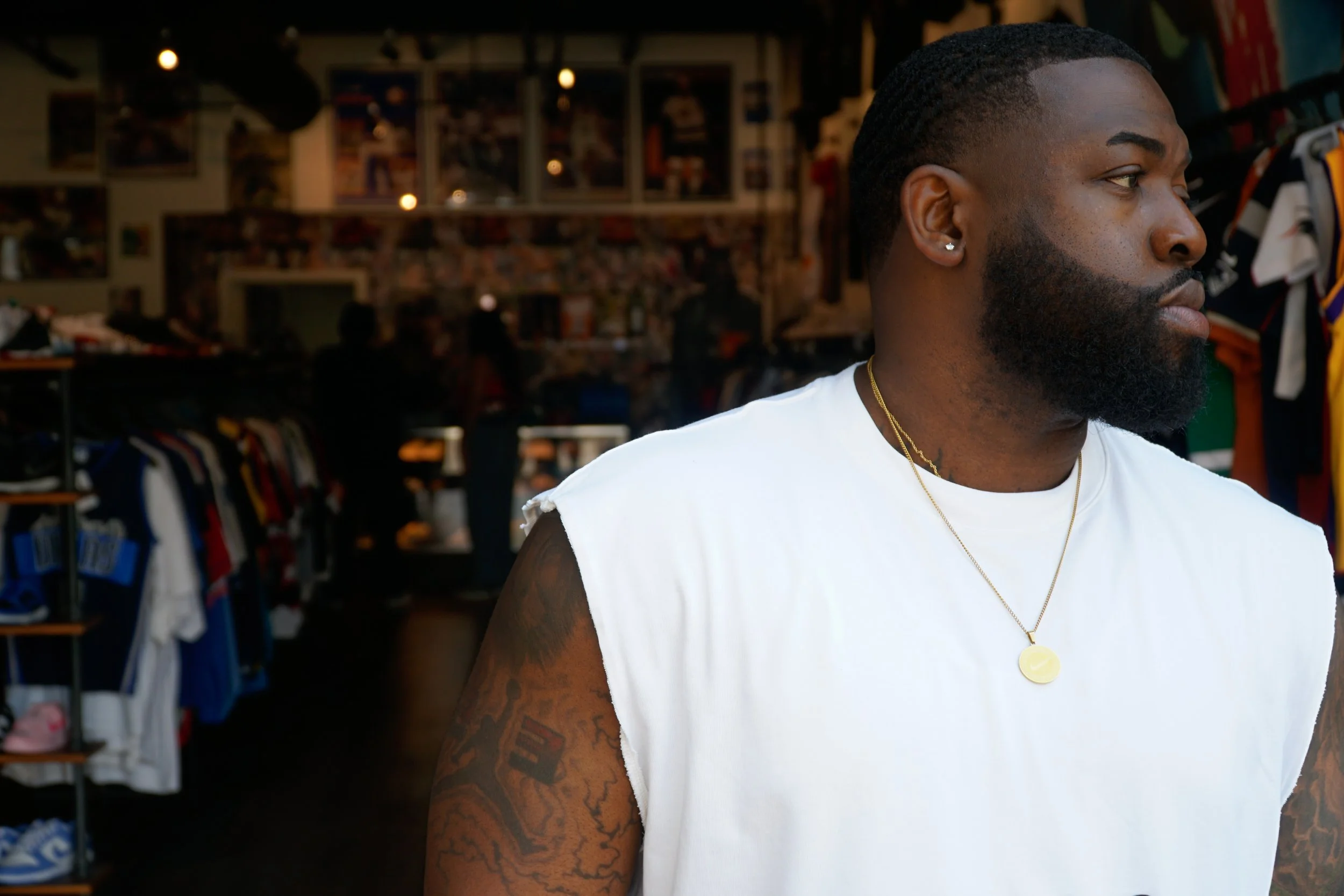 A man with a beard and tattoos on his arm, wearing a sleeveless white shirt and gold necklaces, standing inside a clothing store with shelves of shirts and jackets in the background.