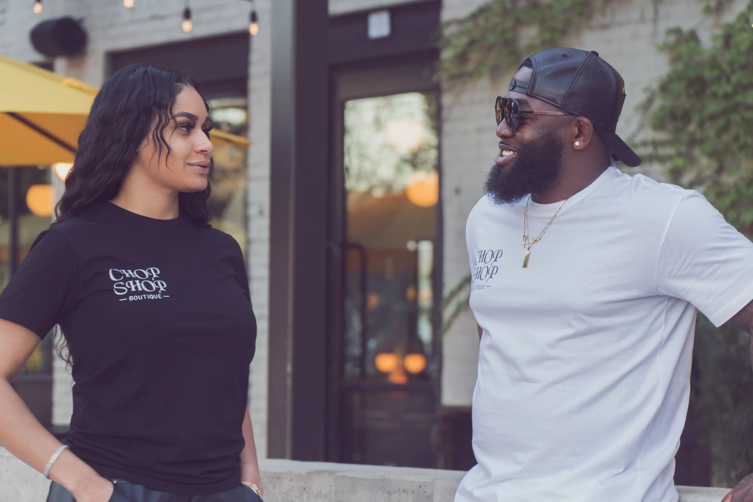 A woman and a man are talking outside a building. The woman is wearing a black T-shirt and the man is wearing a white T-shirt, sunglasses, and a cap turned backward. They are standing near a counter with yellow umbrellas in the background.