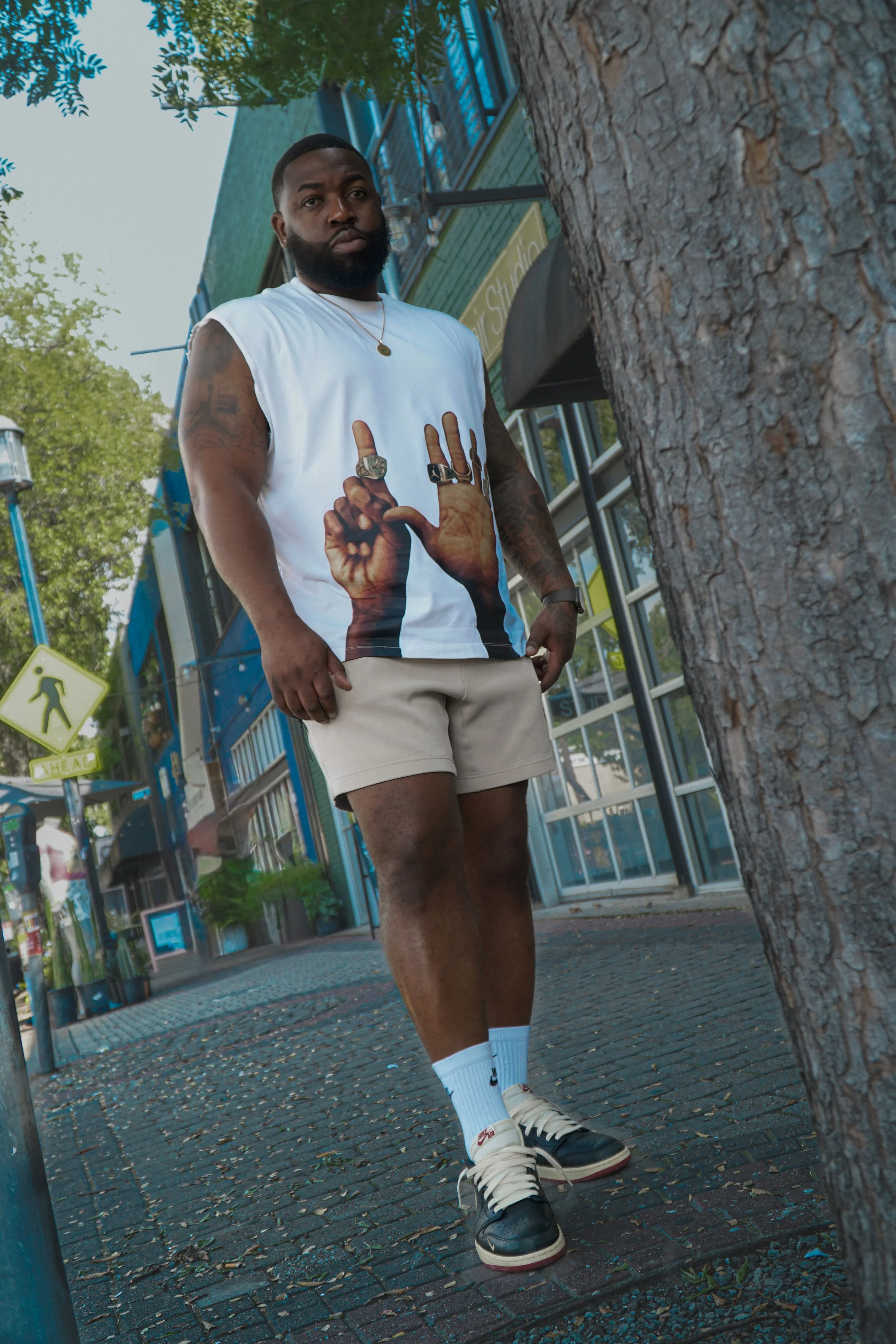 A man with a beard standing on a sidewalk next to a tree, wearing a white sleeveless shirt with handprints and rings, beige shorts, white socks, and black and white sneakers.