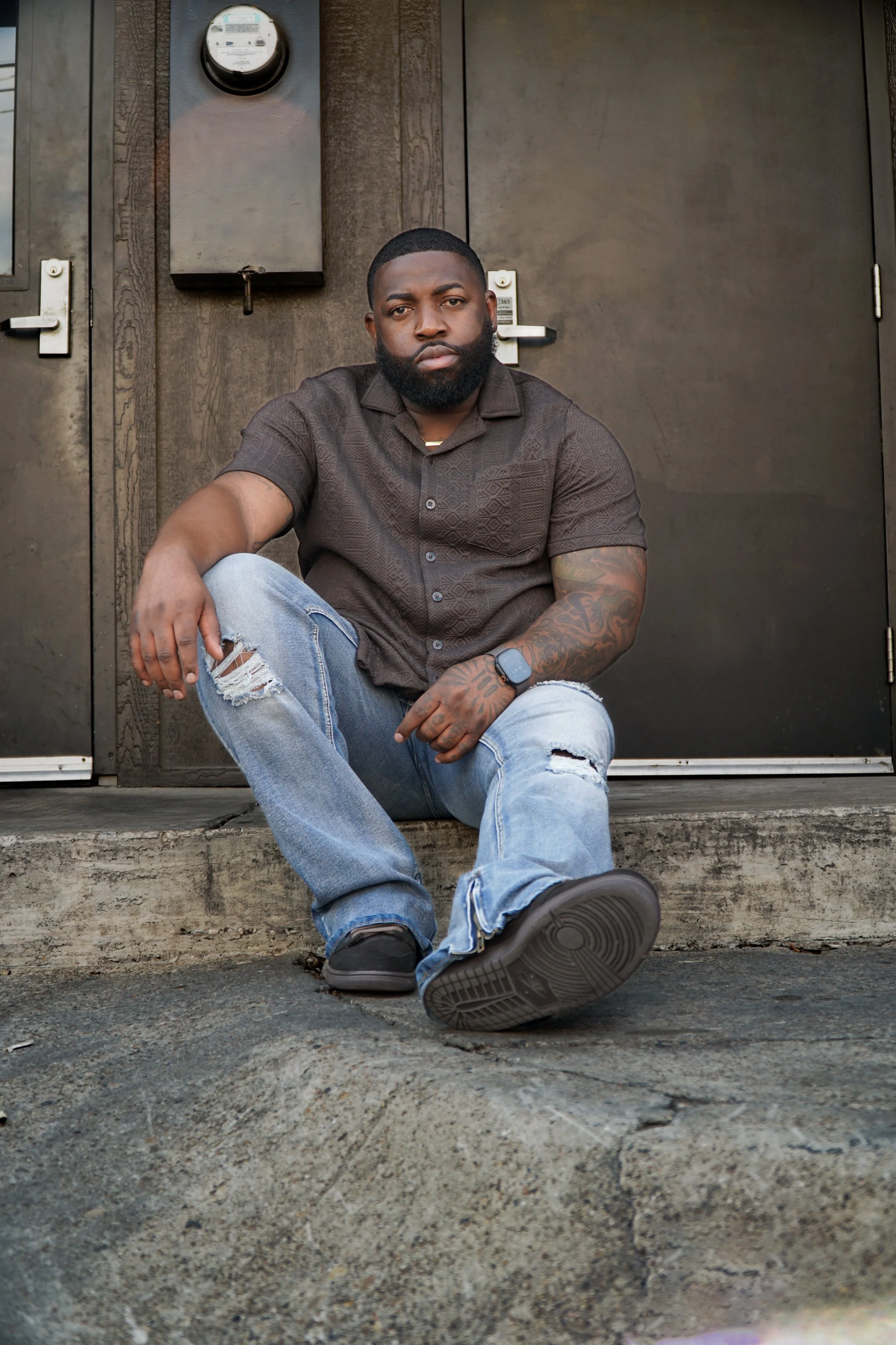 A young man with a beard and tattoos, wearing a dark shirt, ripped jeans, and sneakers, sitting on a concrete step in front of a dark brown door, looking at the camera.