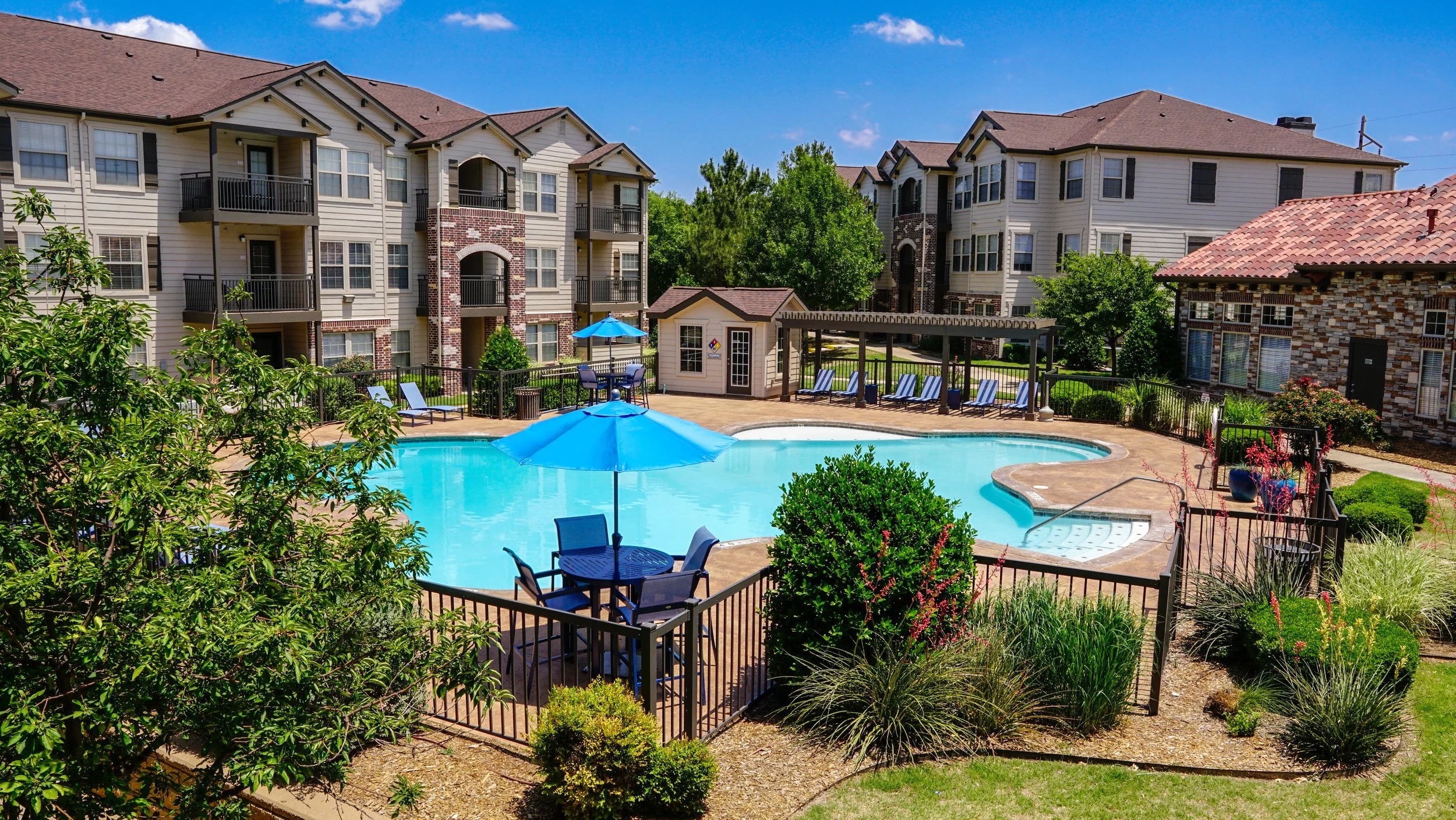 Apartment complex courtyard with swimming pool, lounge chairs, blue umbrellas, landscaped greenery, and surrounding residential buildings.