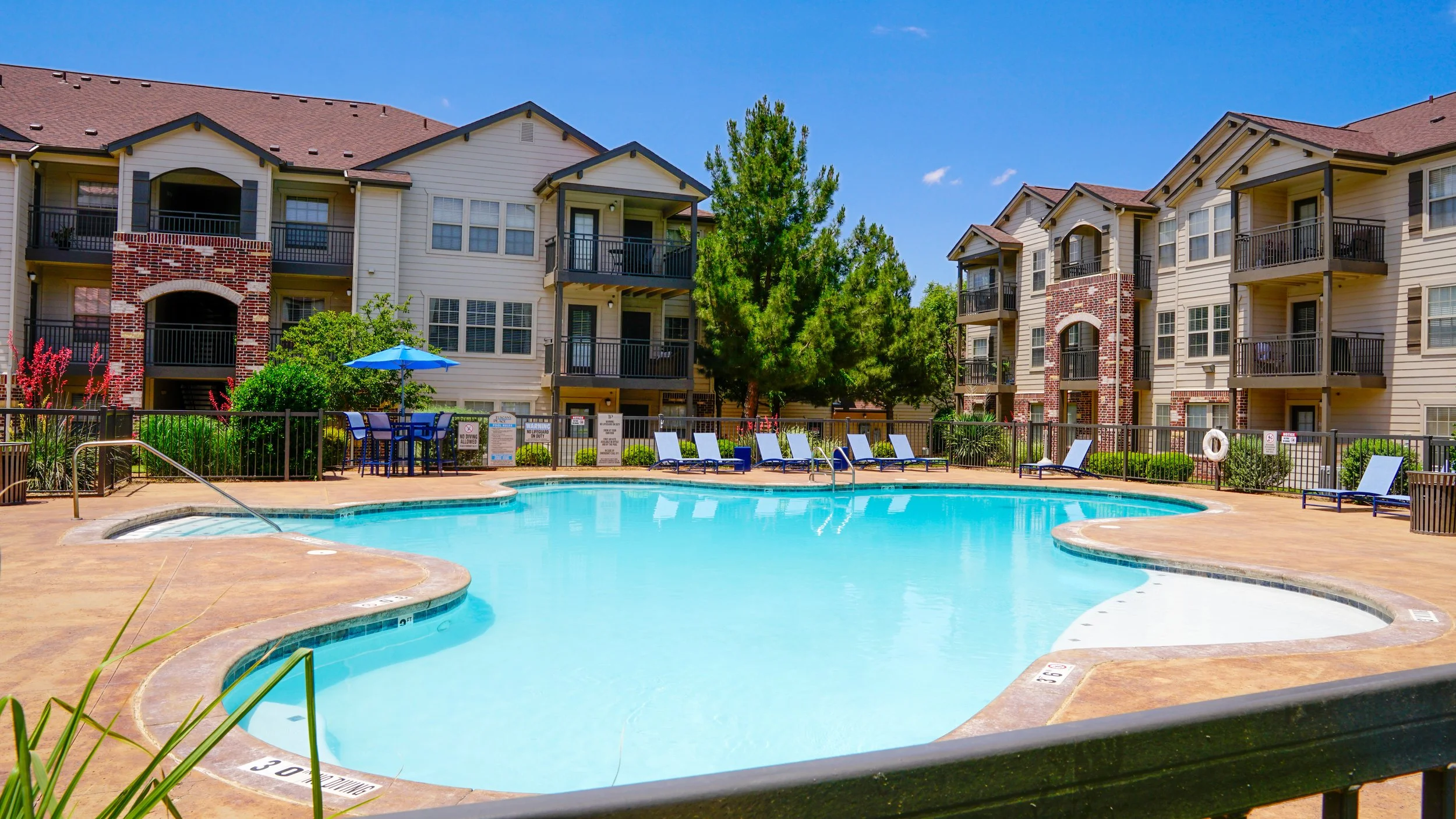 An outdoor swimming pool in an apartment complex with lounge chairs, a table with an umbrella, surrounded by a black fence, with multistory apartment buildings and green trees in the background.