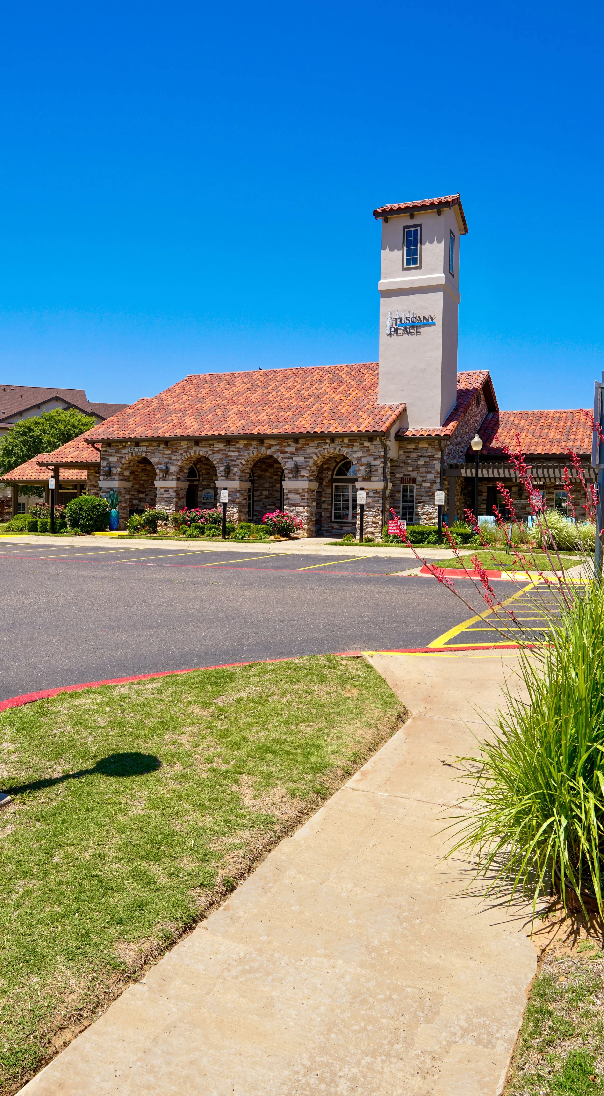 A building named Toscana Place with a brick facade, tiled roof, and a tall white tower sign, surrounded by a parking lot and landscaped garden with pink flowers under a clear blue sky.