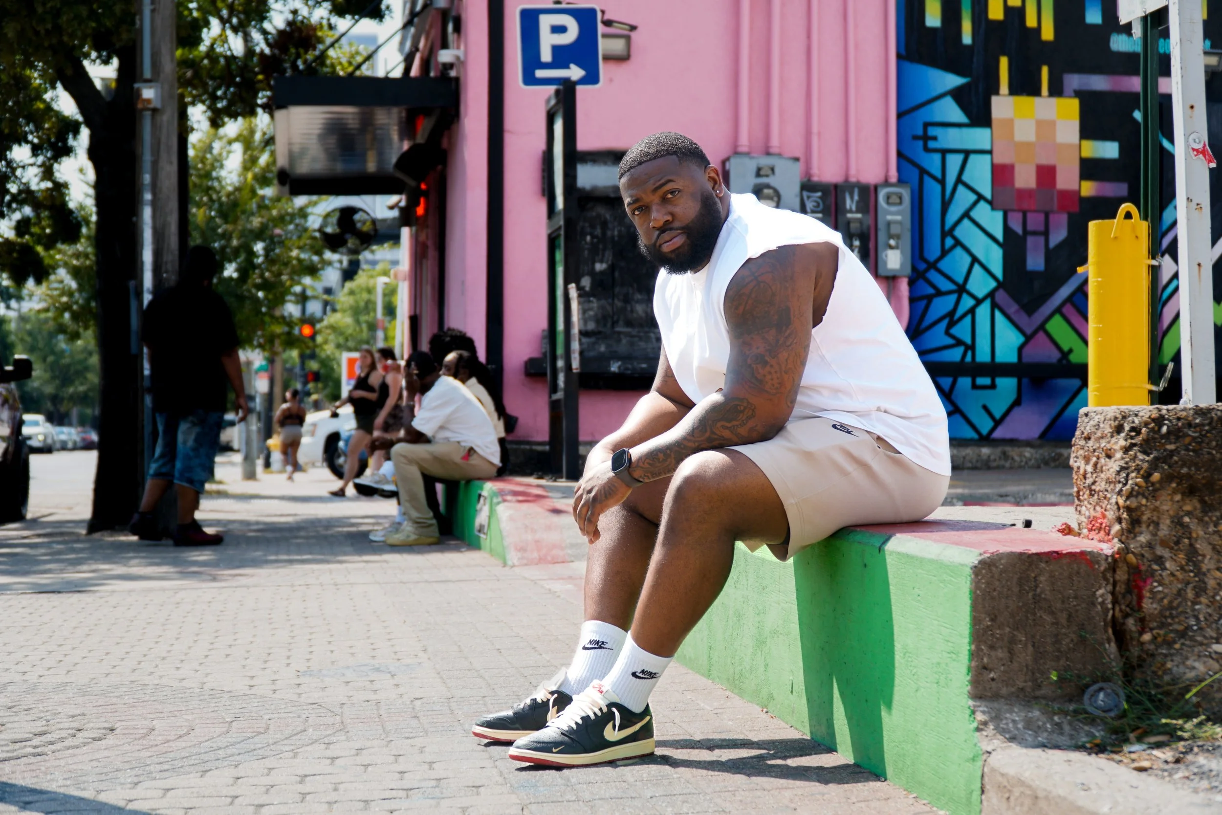 A man with a beard and tattoos sitting on a colorful sidewalk bench in an urban area with other people sitting and standing nearby, against a backdrop of pink and mural art on buildings.