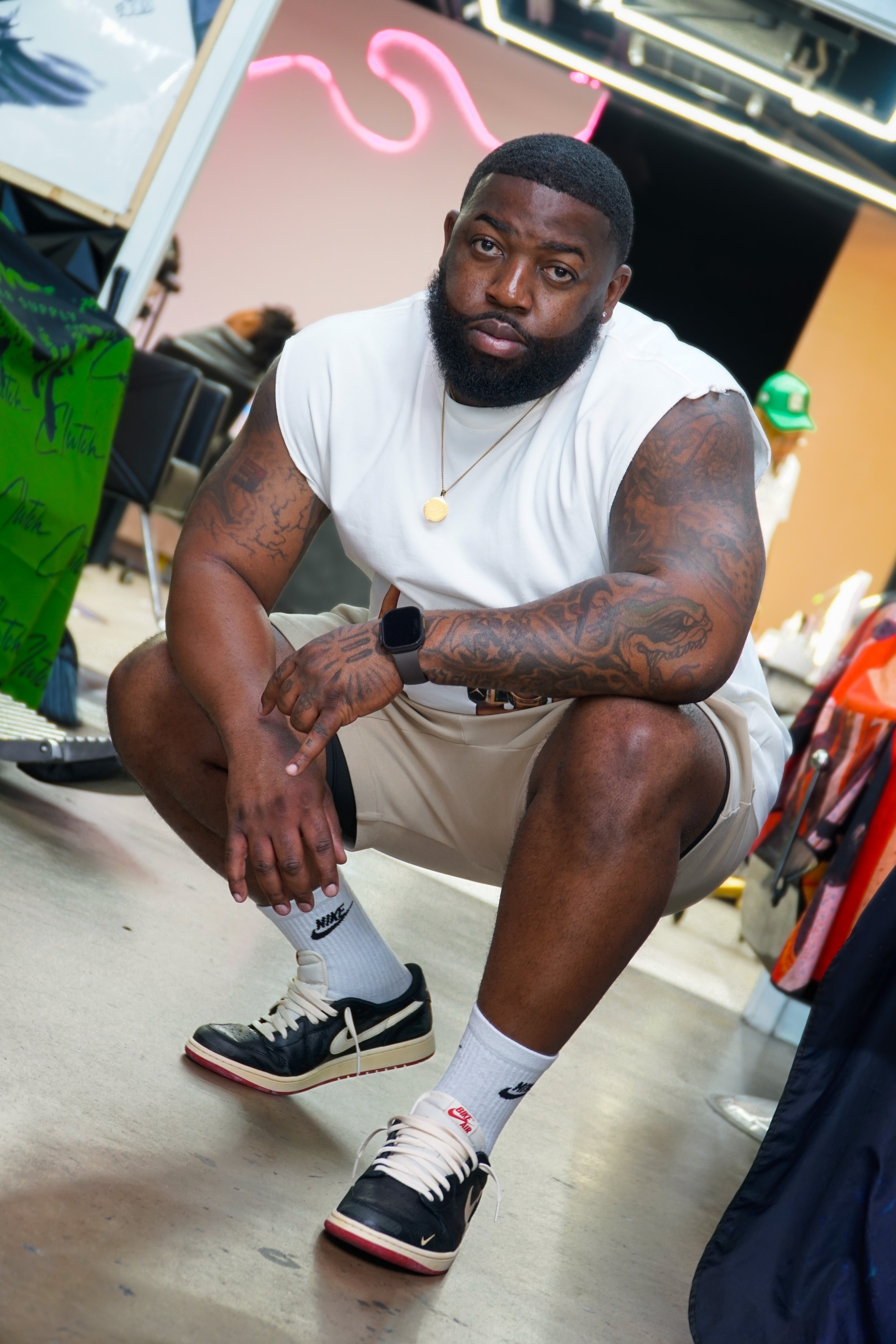 A man with a beard and tattoos squatting indoors. He is wearing a white sleeveless shirt, beige shorts, white Nike socks, and Nike sneakers. There is a neon pink sign in the background and various items around him.