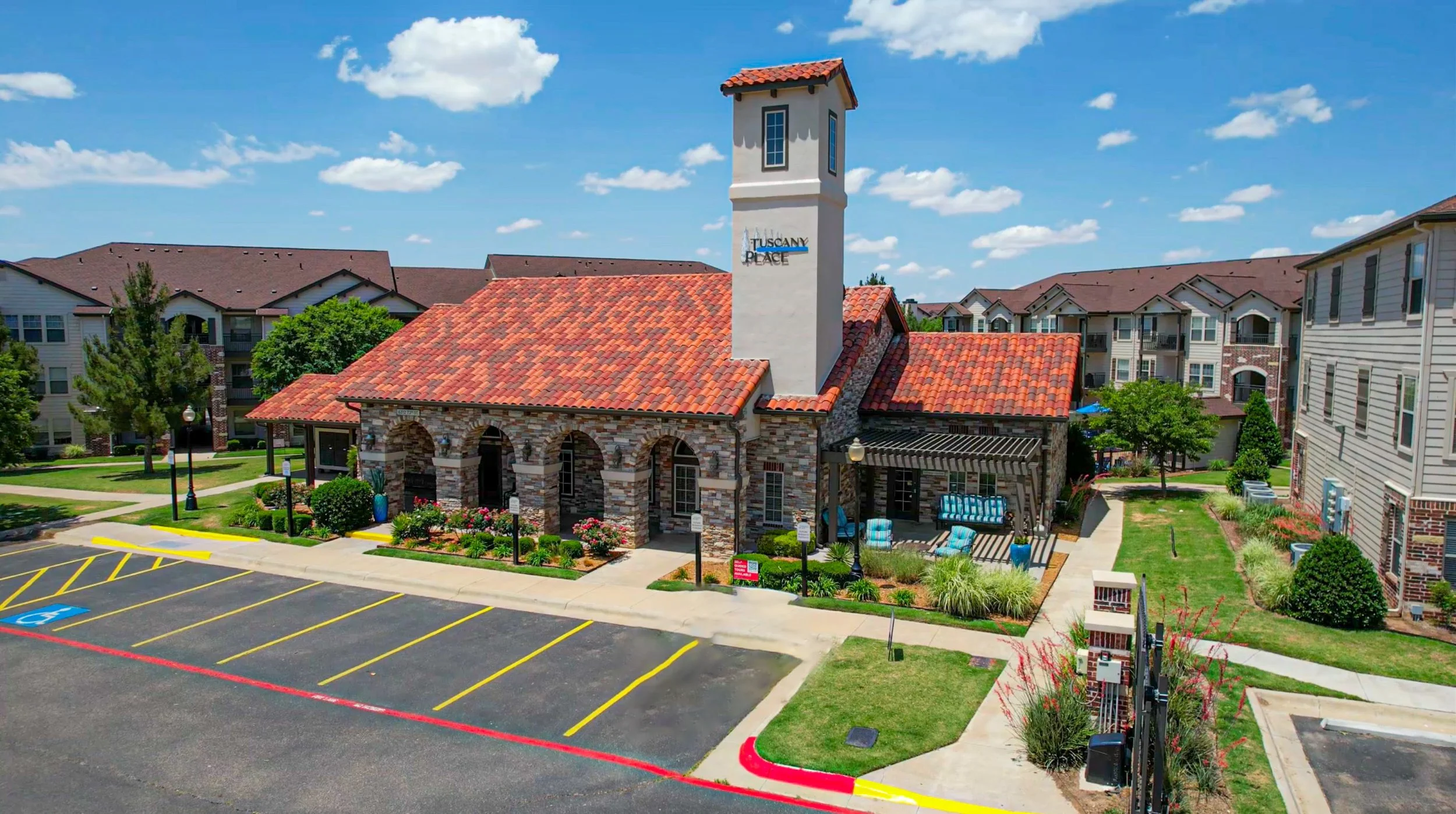 A small building with a red-tiled roof, stone archway entrances, and a tall white tower with windows, labeled 'Tuscany Place,' surrounded by a parking lot, green lawns, and neighboring residential buildings on a sunny day.