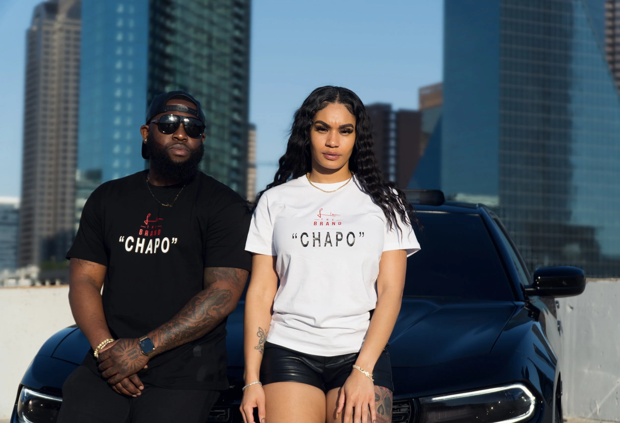 A man and woman standing in front of a black car with a cityscape background, both wearing T-shirts with the text 'Chapo' and posing confidently.