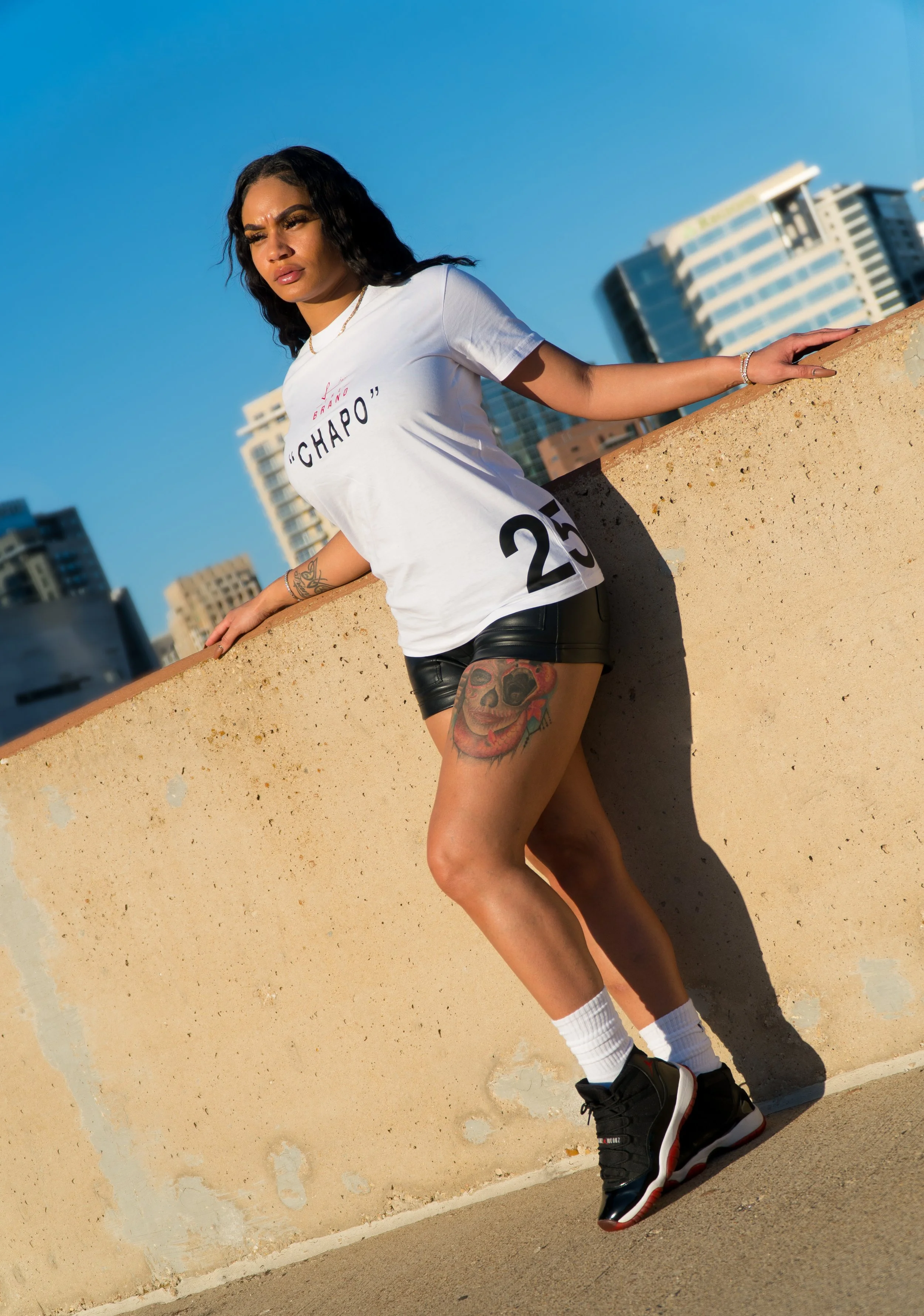 A woman standing on a rooftop balcony against a cityscape background, wearing a white T-shirt, black shorts, white socks, and black sneakers.