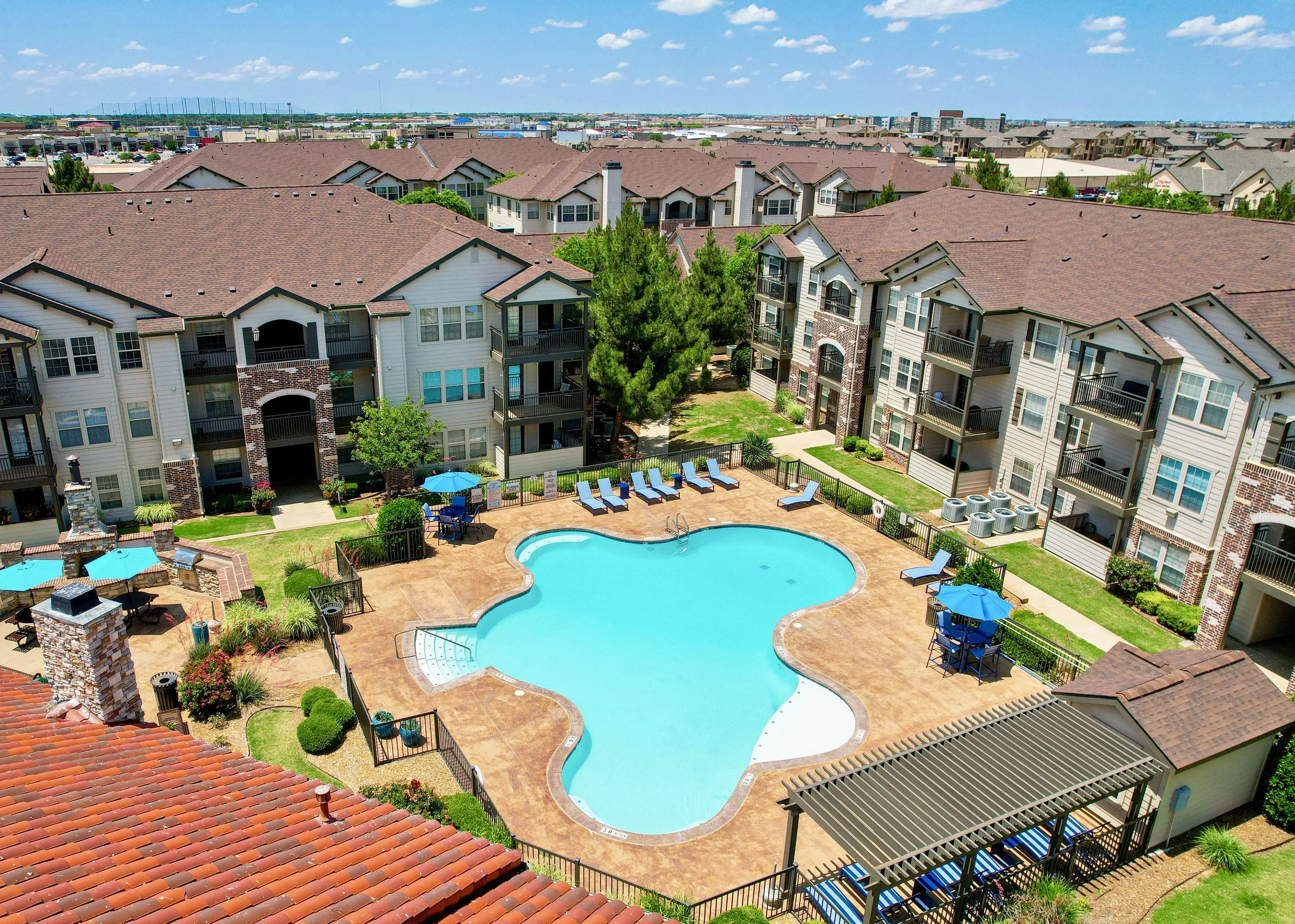 View of a residential apartment complex with a central outdoor swimming pool, surrounded by lounge chairs, umbrellas, and landscaped greenery under a partly cloudy sky.