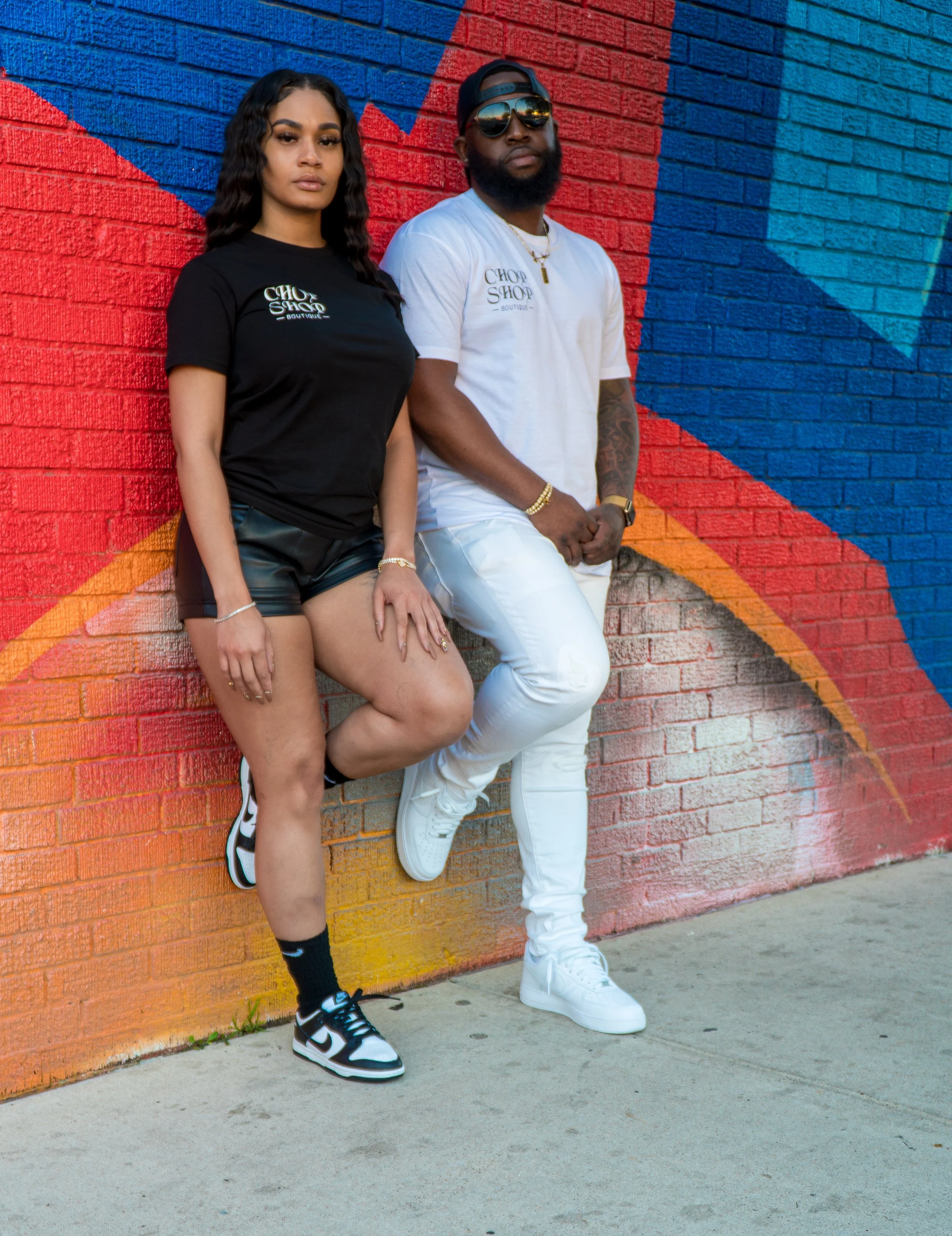 Two young adults, a woman and a man, stand against a colorful, mural-painted brick wall. The woman is wearing a black T-shirt, black shorts, and sneakers, with long wavy hair. The man is wearing a white T-shirt, white pants, and white sneakers, with 