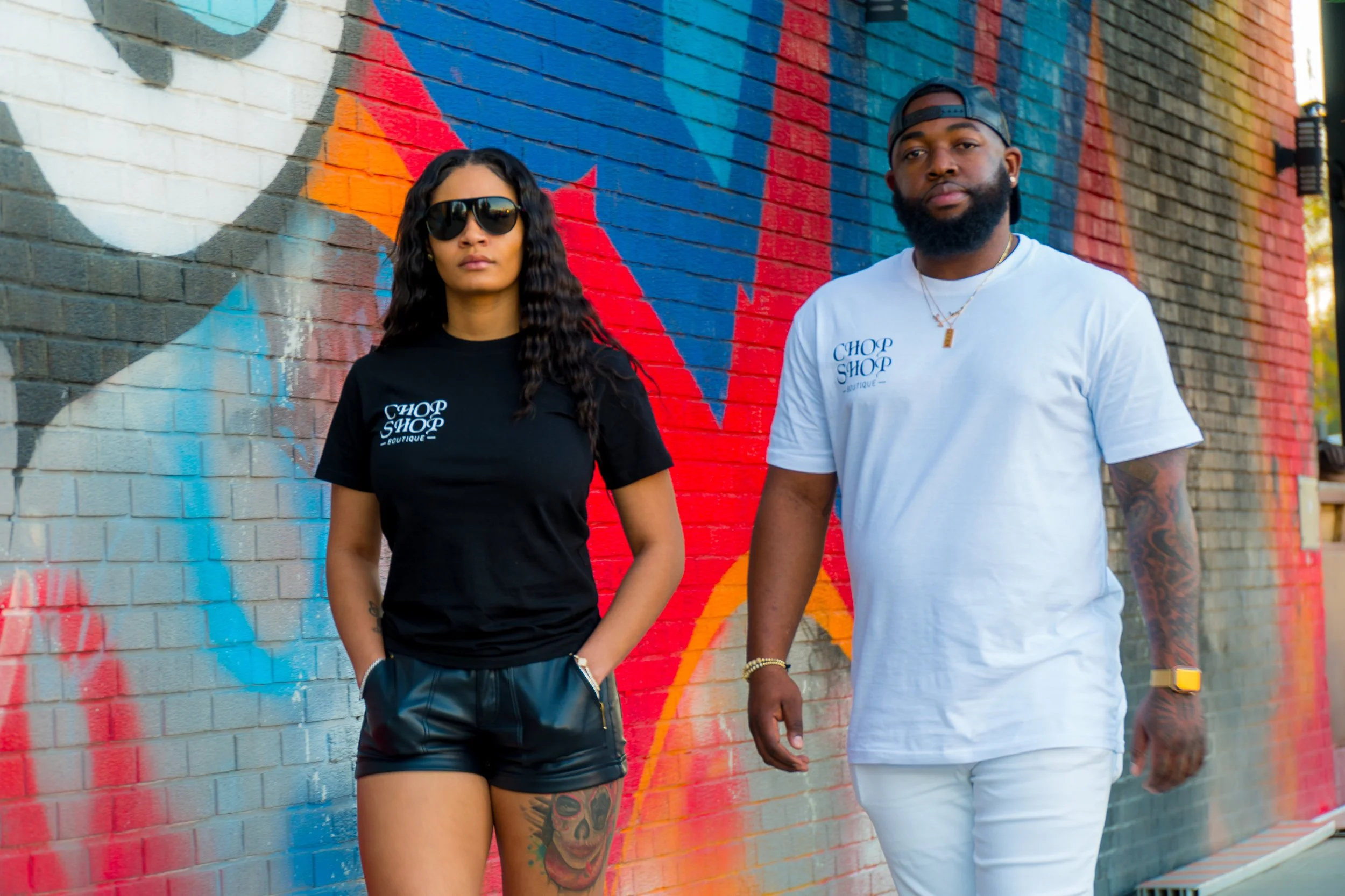 Two young adults, a woman and a man, stand in front of a colorful graffiti mural on a brick wall. The woman wears black sunglasses, a black T-shirt, and black shorts. The man wears a white T-shirt, white pants, and a backwards black cap. Both have ta