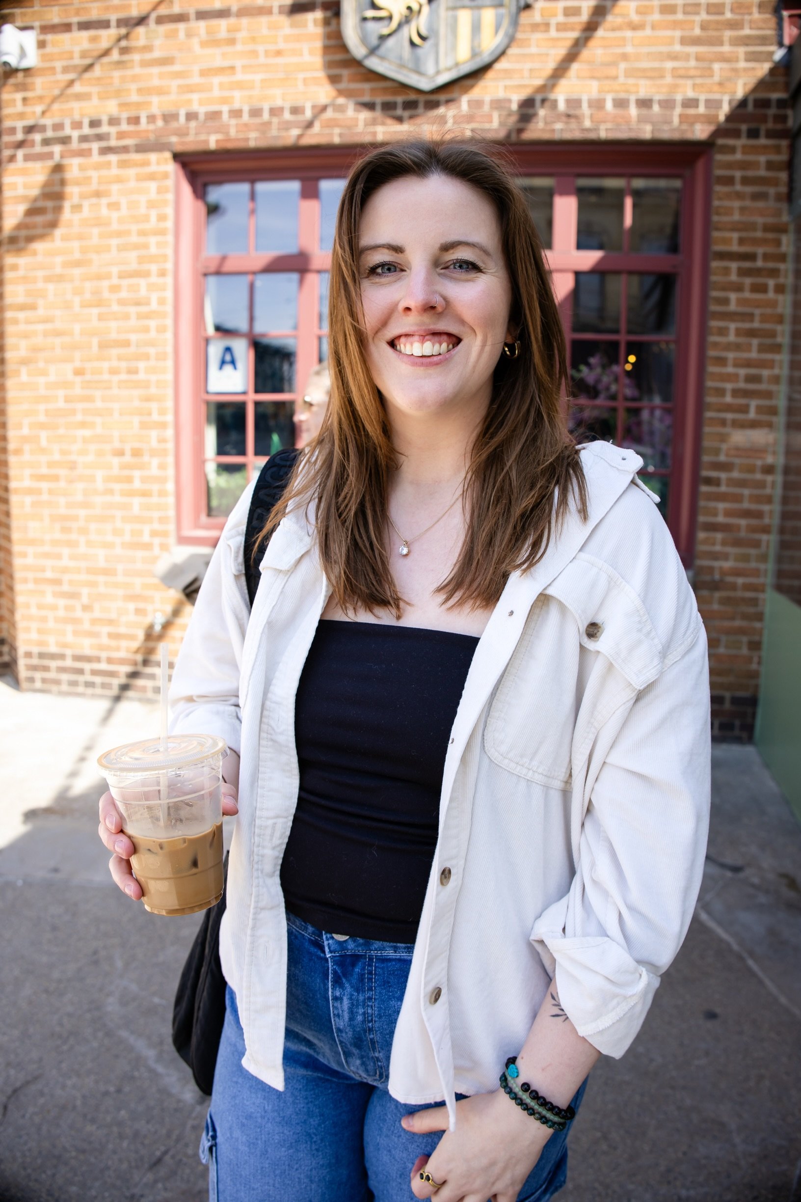 A young woman with brown hair, smiling, wearing a white jacket over a black top, holding a plastic cup of iced coffee, standing outside in front of a brick building with large windows.