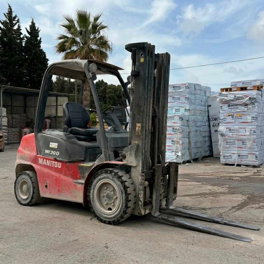 Carrello elevatore Manitou rosso con forche frontali, parcheggiato in un piazzale di materiali edili, con pallet di sacchi impilati e alberi sullo sfondo.