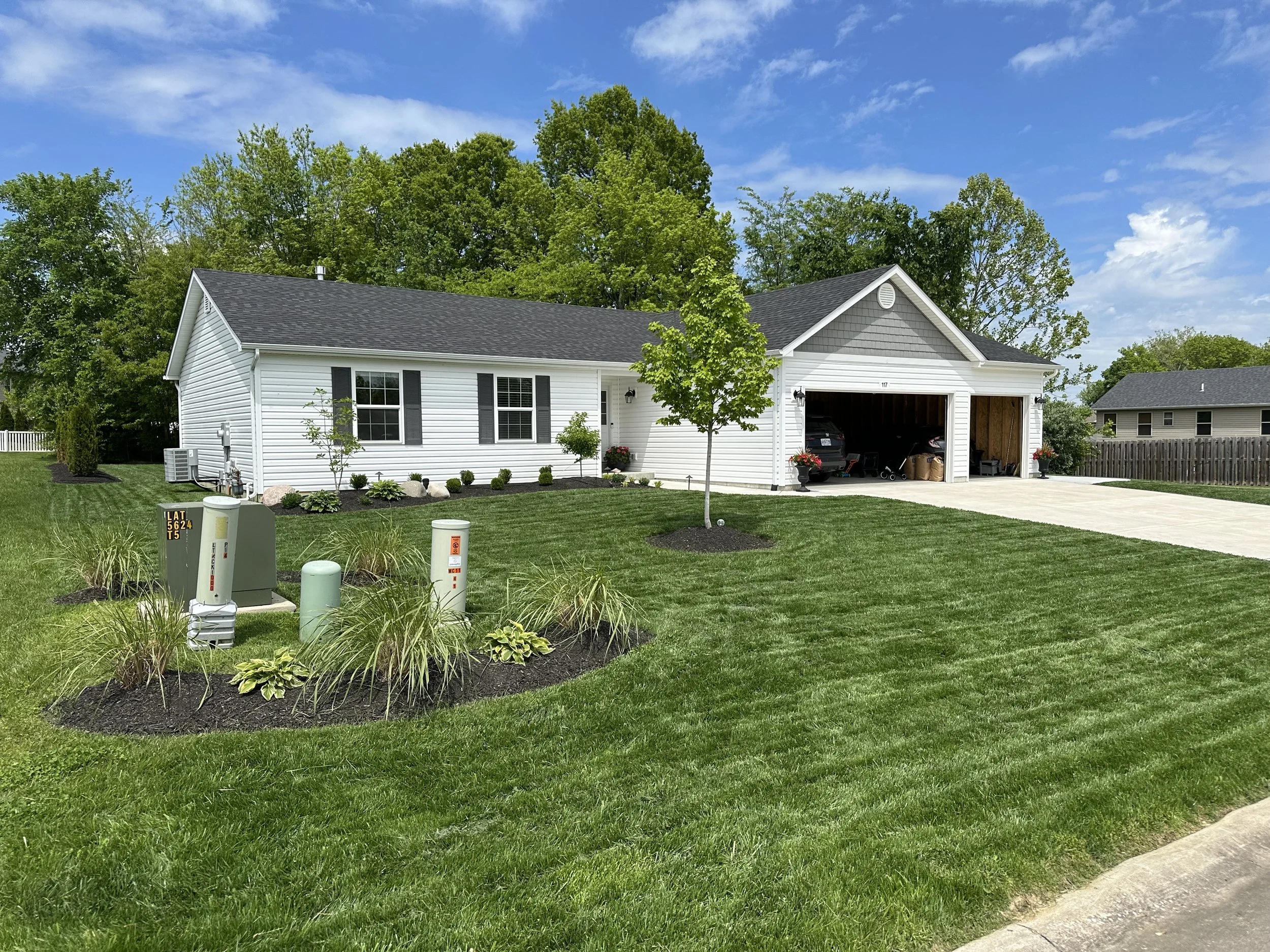 White single-story house with attached garage, green lawn, small tree, landscaped flower beds, and a concrete driveway on a sunny day.