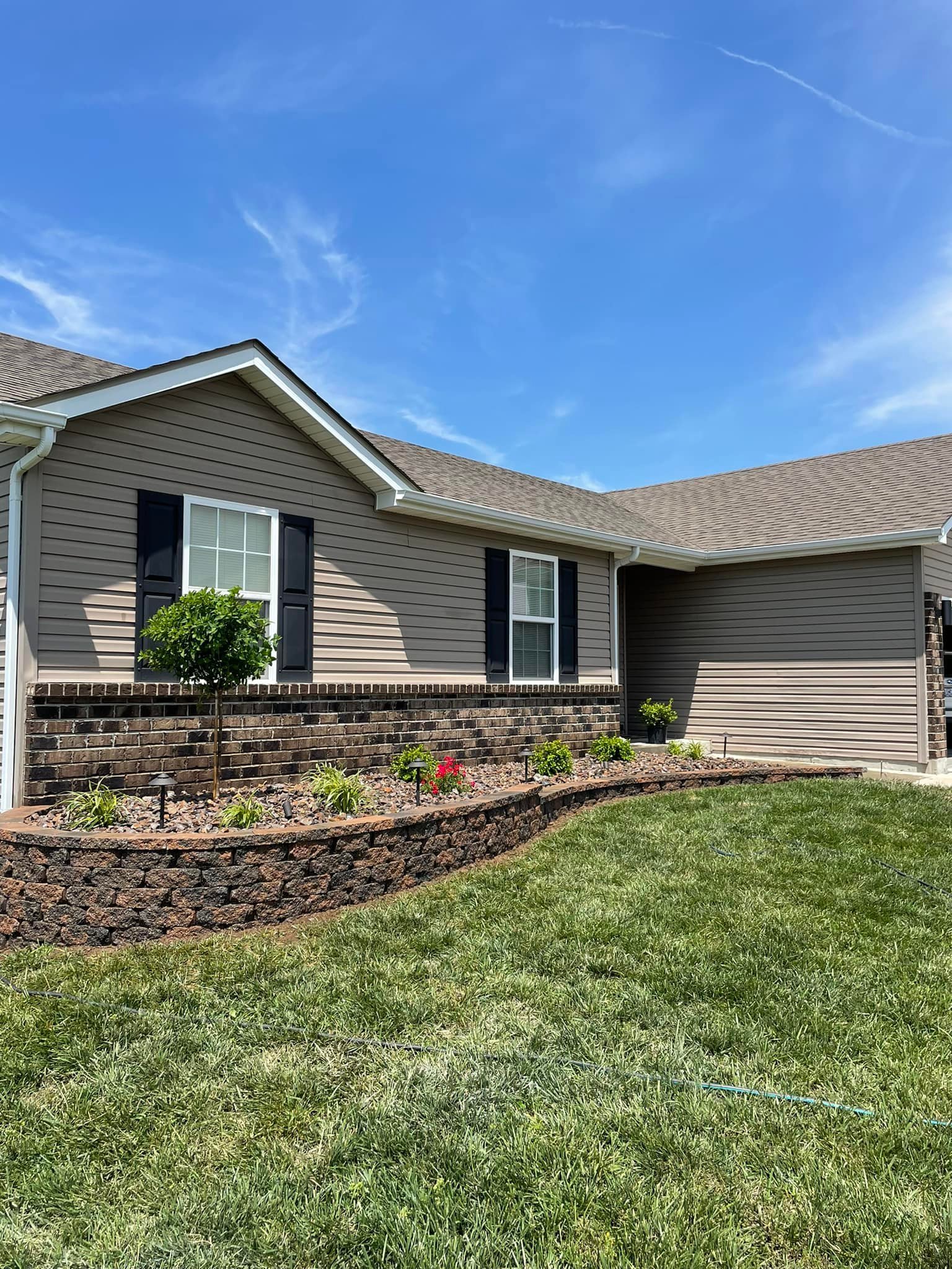 Front yard of a house with a small garden bed bordered by decorative stone, a green lawn, and beige siding with white and black window shutters. Clear blue sky in the background.