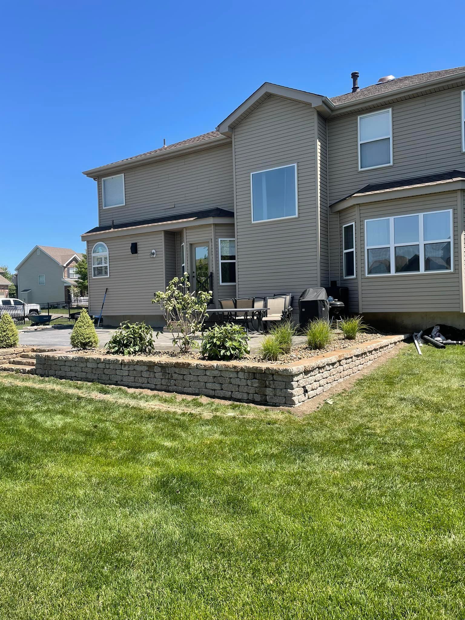 A two-story house with beige siding and multiple large windows overlooking a well-maintained backyard. The backyard features a raised brick garden bed with plants and bushes, and a patio area with outdoor furniture and a barbecue grill. The grass is green, and the sky is clear with no clouds.
