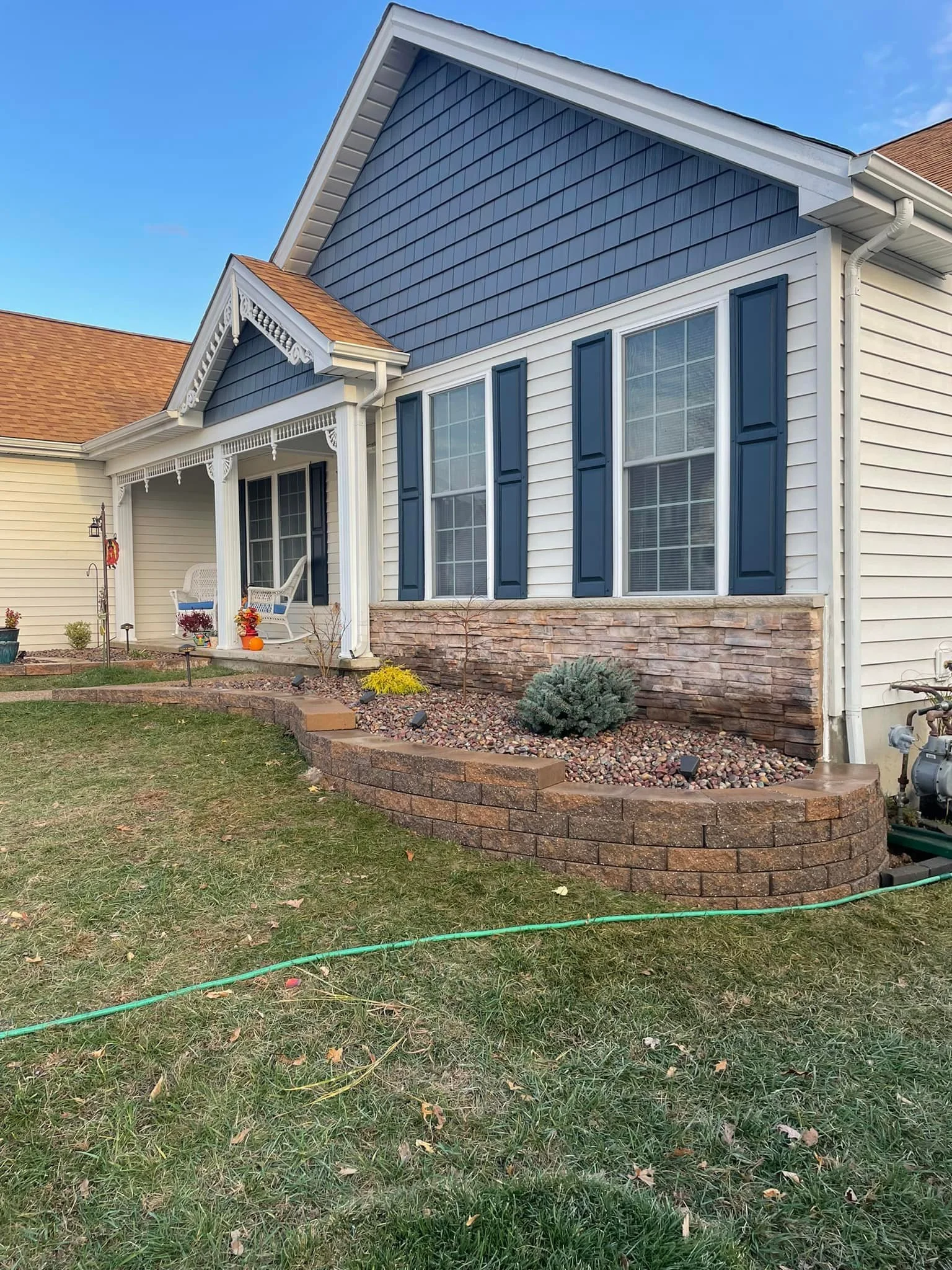 Front yard and porch of a house with a blue and white exterior, brown brick landscaping wall, and a small porch with white chairs and decorations.