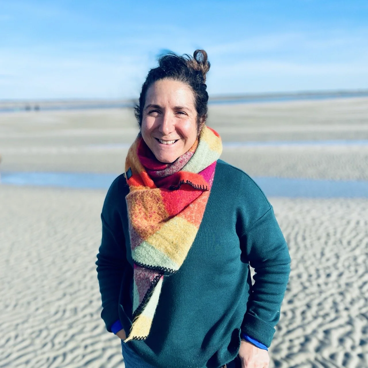 Une femme souriante porte un foulard coloré sur une plage sablonneuse avec un ciel bleu et de l'eau en arrière-plan.