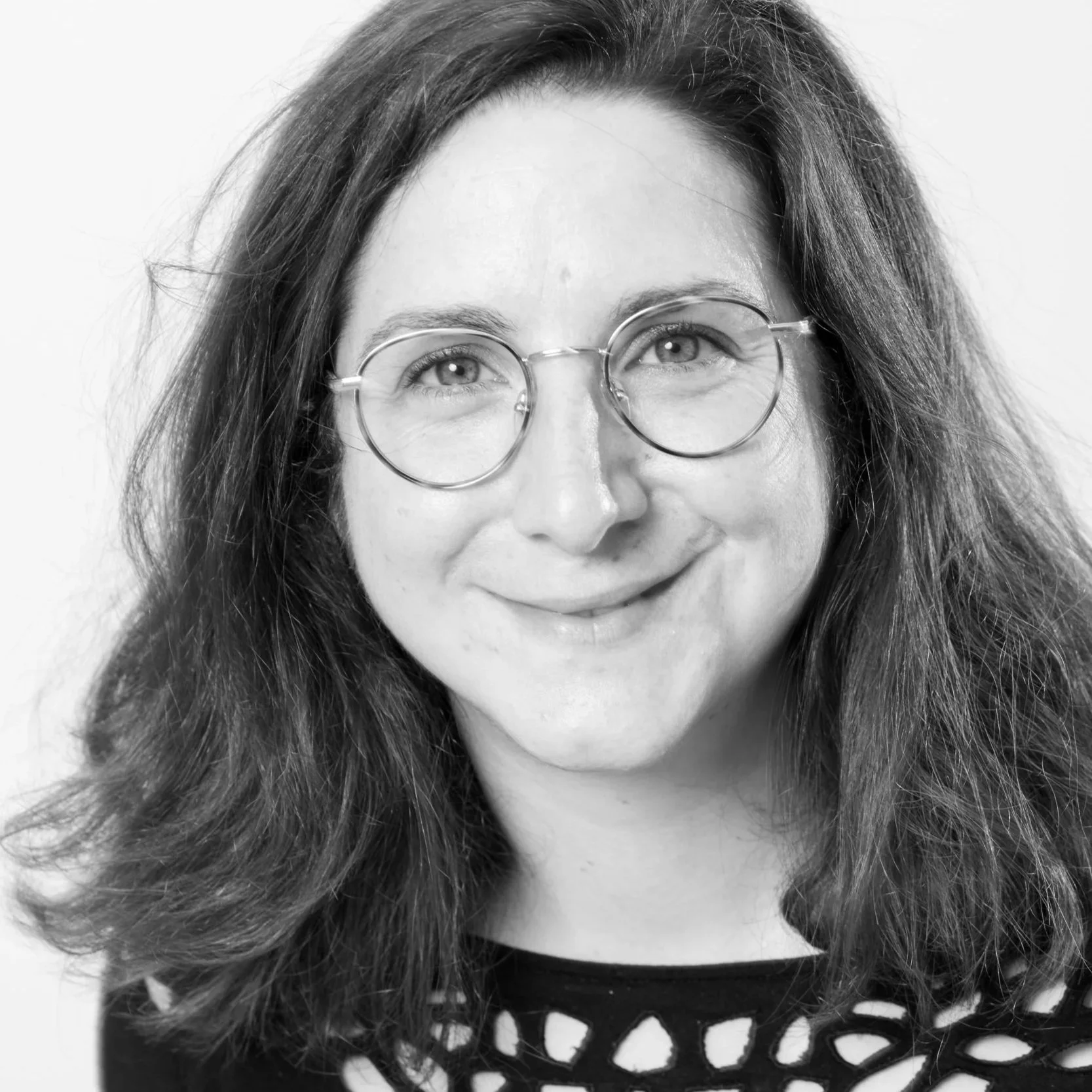 Portrait en noir et blanc d'une femme avec des lunettes rondes, souriante, aux cheveux longs et ondulés, portant un haut avec un motif lacé.