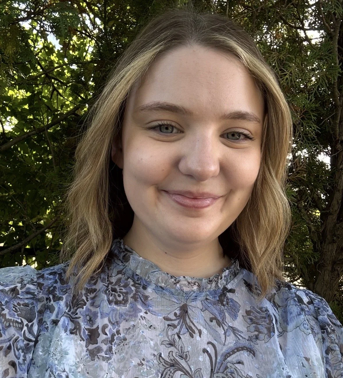A young woman with shoulder-length wavy hair and light skin is smiling outdoors in front of green foliage, wearing a light-colored blouse with a floral pattern.