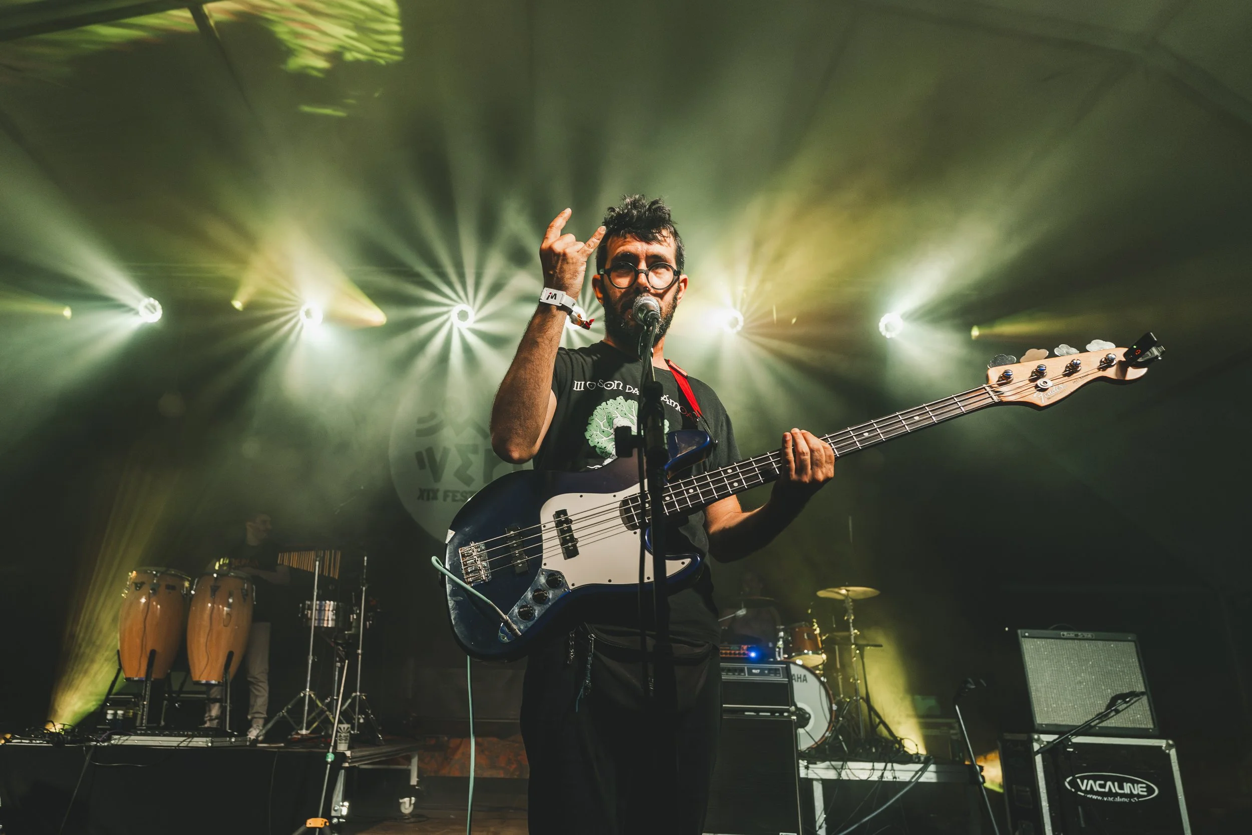 Hombre tocando el bajo en un escenario con luces de colores y instrumentos musicales en el fondo.