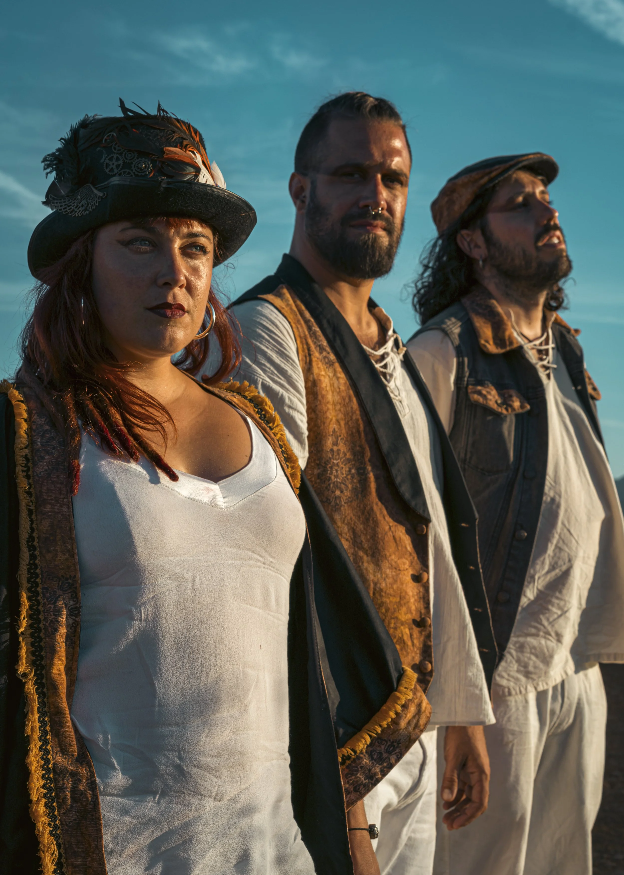 Tres personas con vestimenta de estilo folclórico posando al atardecer, con cielo azul y nubes en el fondo.