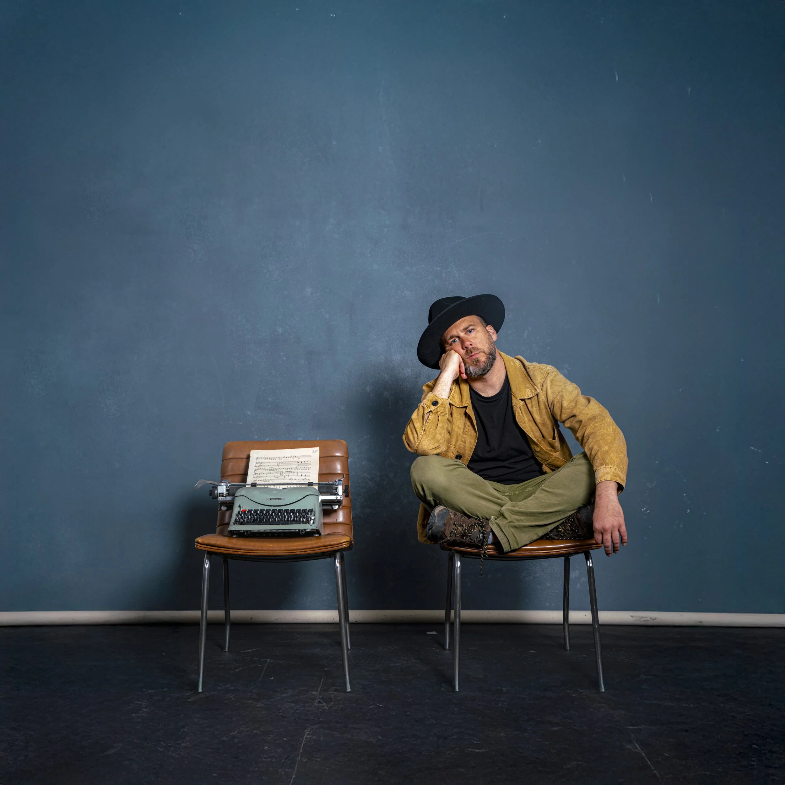 Un hombre sentado en una silla con una máquina de escribir frente a él, en un fondo de pared azul. La máquina de escribir tiene una hoja con partituras musicales.
