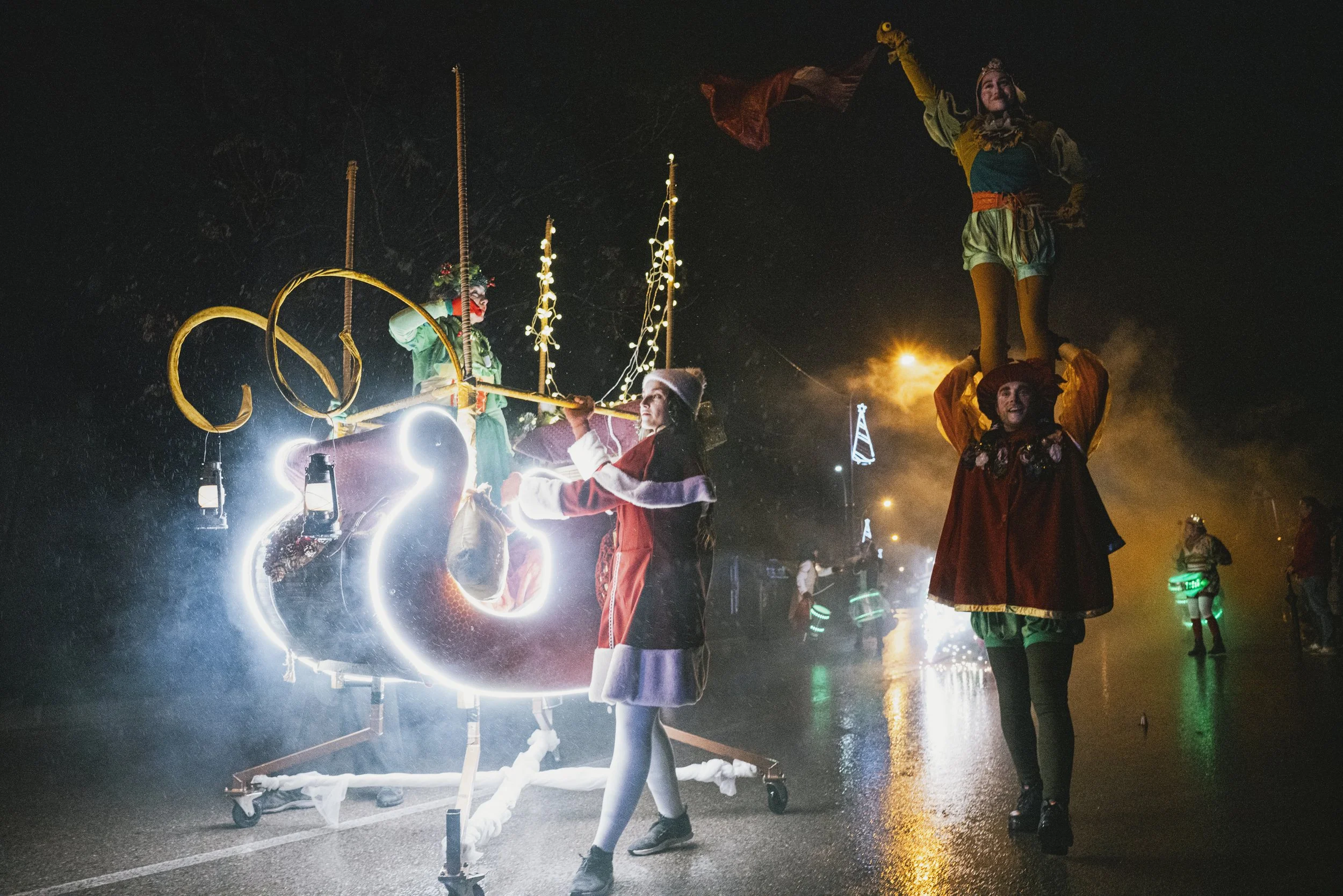 Personajes disfrazados de Navidad realizando un espectáculo en la calle en la noche lluviosa, uno de ellos soplando un caño decorado con luces y otros equilibrando a una persona en la cabeza.