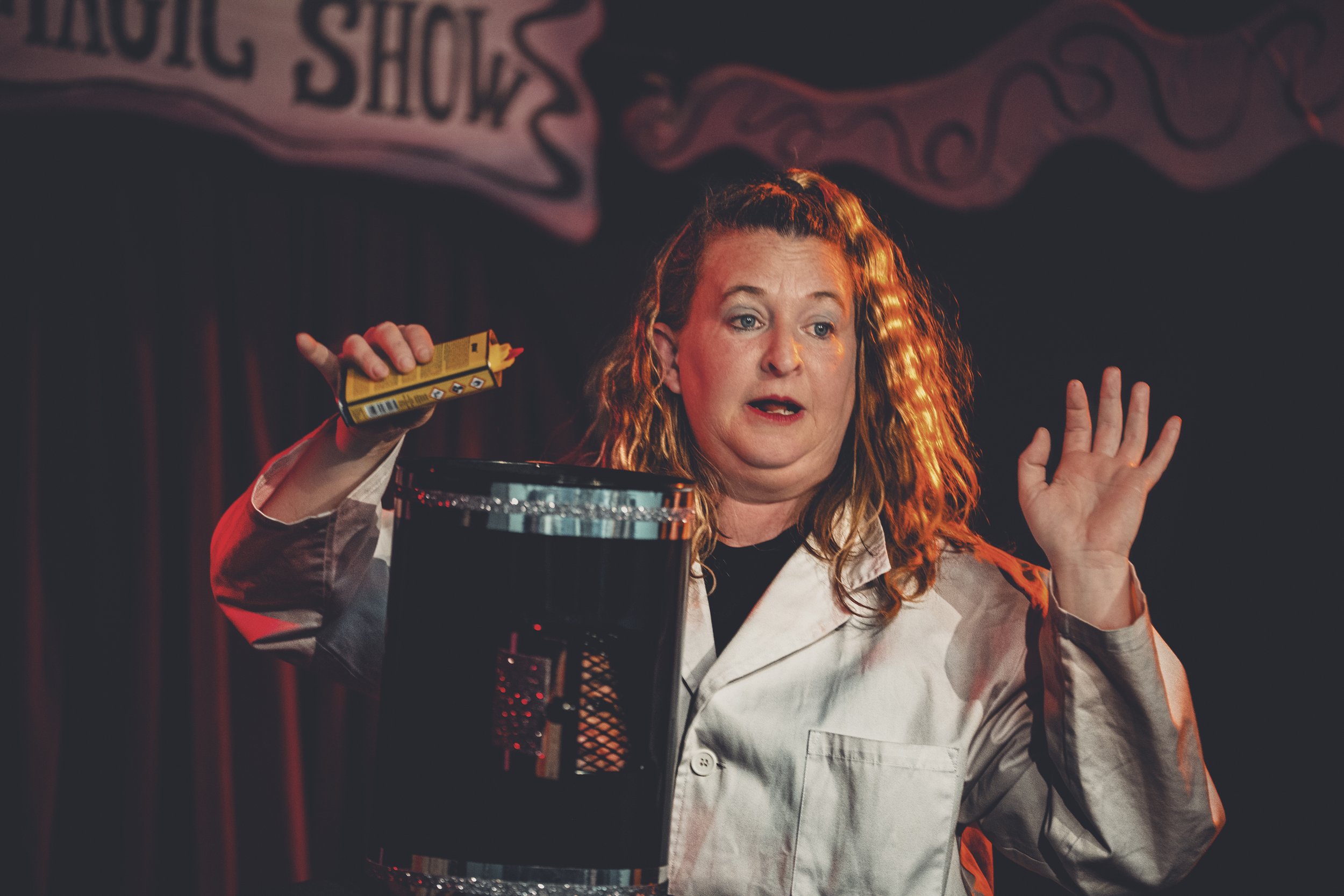 Una mujer actuando en un espectáculo de comedia, sosteniendo una caja de cerillas en una mano y levantando la otra mano con expresión de sorpresa o énfasis, en un escenario con cortinas oscuras y un cartel que dice 'Magic Show'.