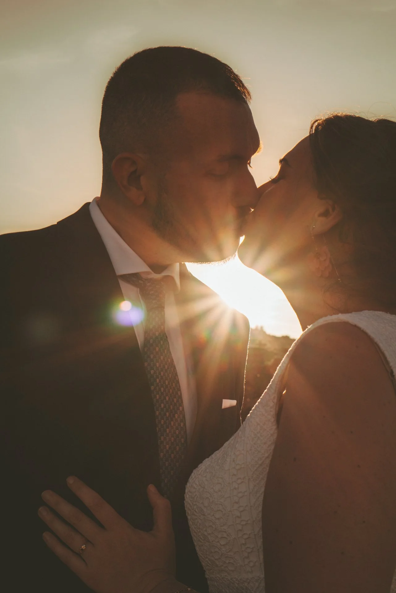 Una pareja en boda besándose al atardecer con sol brillante detrás.