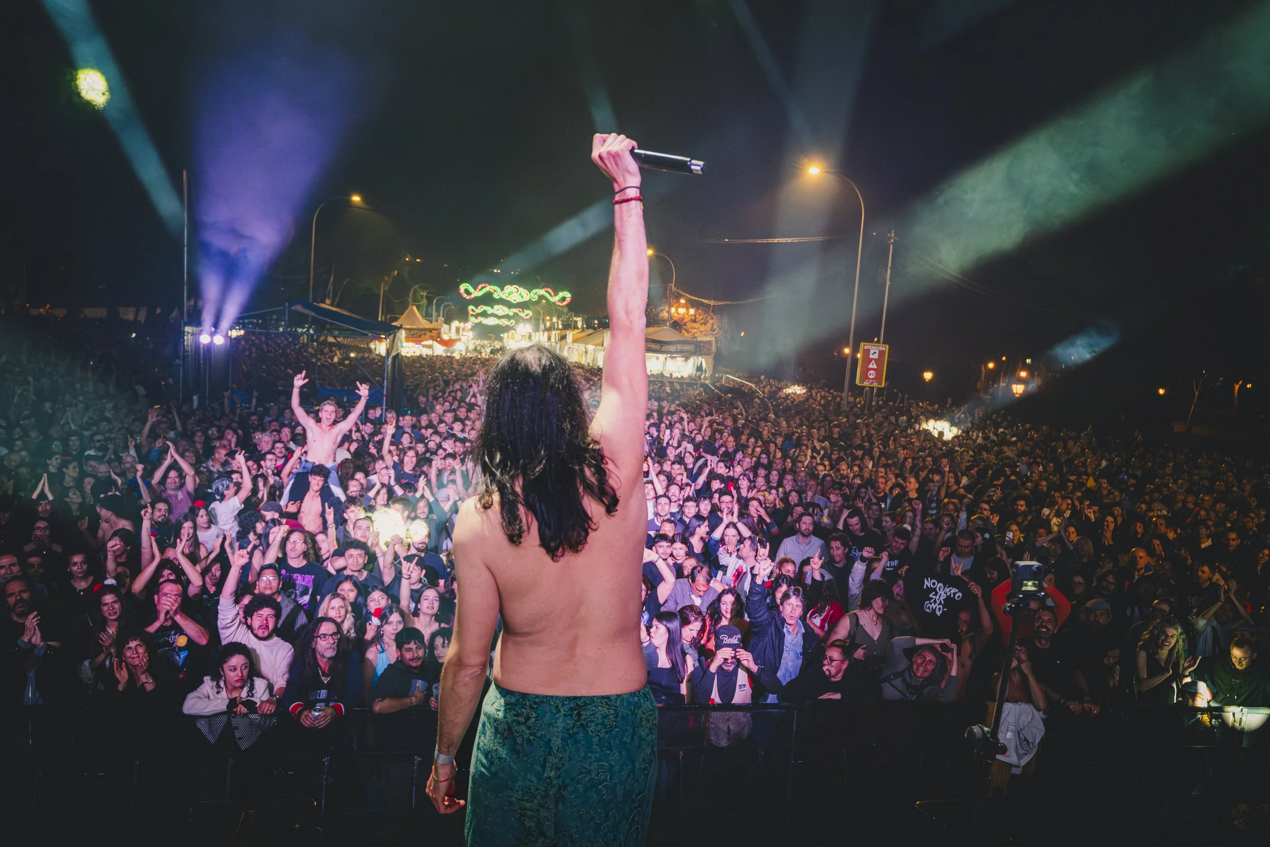 Persona en el escenario durante un concierto o festival, vista desde atrás, sosteniendo un micrófono en alto, con una multitud de espectadores frente a ella, luces y colores vivos en el fondo.