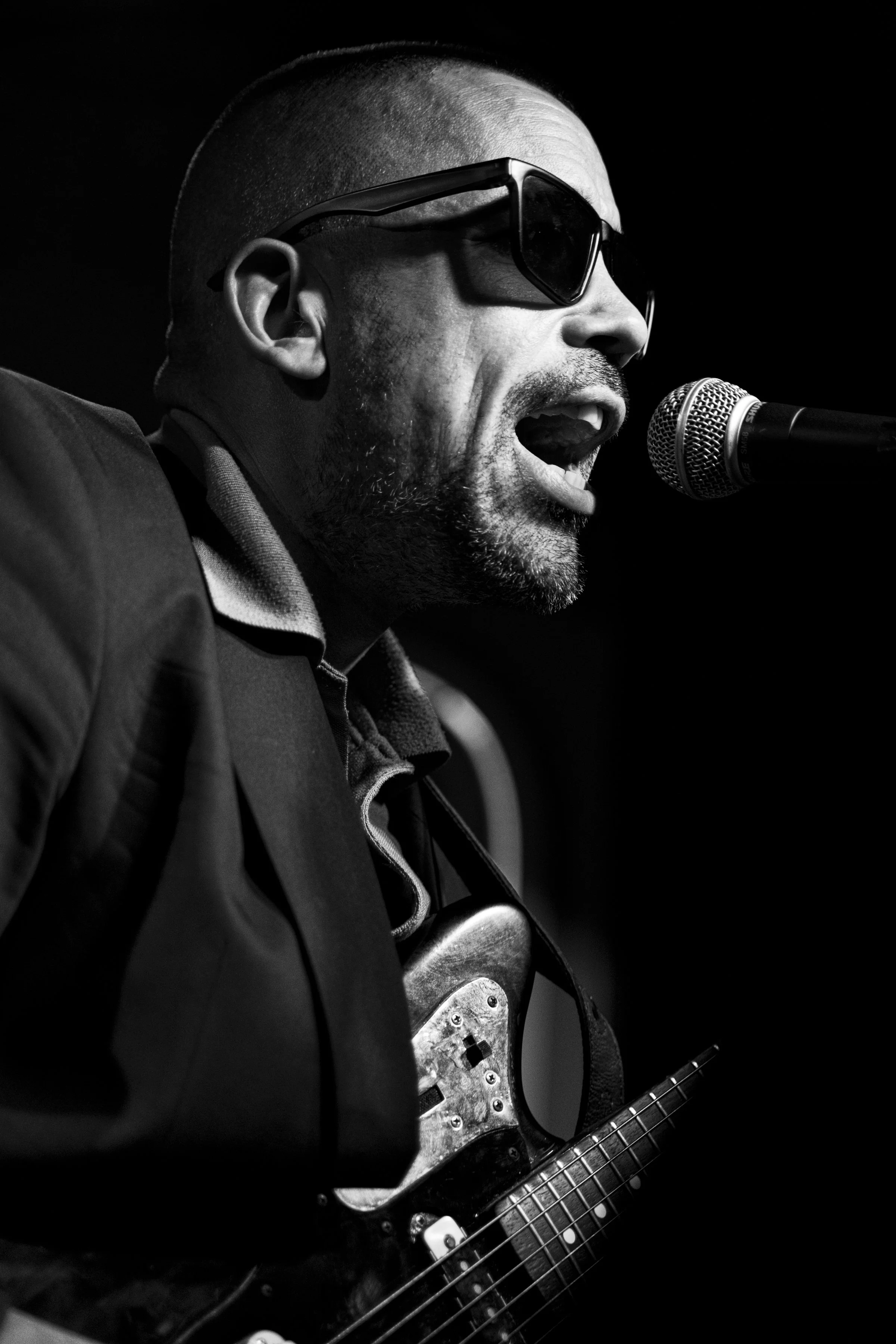 Hombre tocando guitarra eléctrica y cantando frente a un micrófono en un escenario, con gafas oscuras, en una foto en blanco y negro.