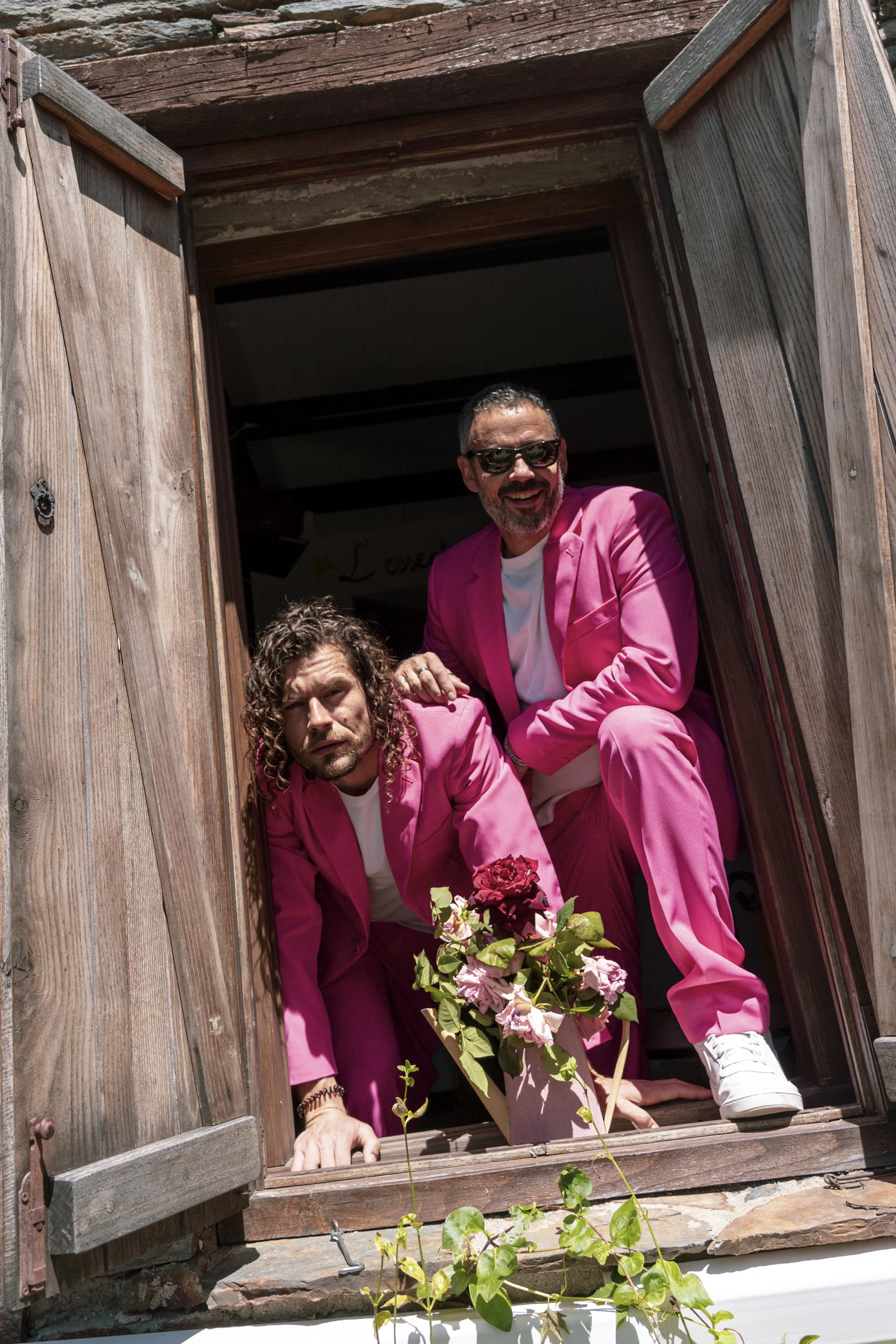 Dos hombres con trajes rosados, uno con barba y lentes oscuros, están asomados desde una ventana de madera, también llevan camisetas blancas, hay plantas con flores en un jarrón en el alféizar de la ventana.