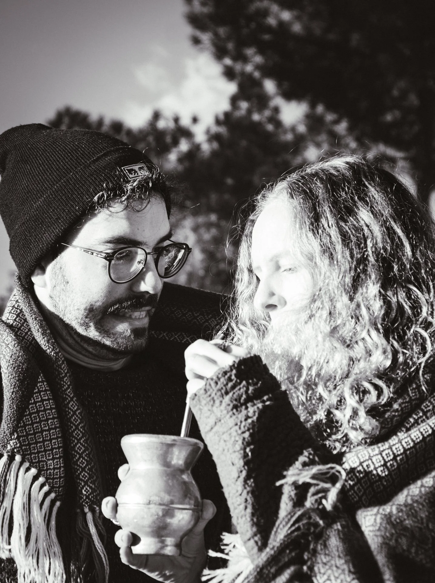 Dos personas en exteriores, una sosteniendo una tradicional mate con bombilla, ella tiene cabello rizado y parece estar a punto de beber, él lleva gafas y gorro, compartiendo un momento en un día soleado.