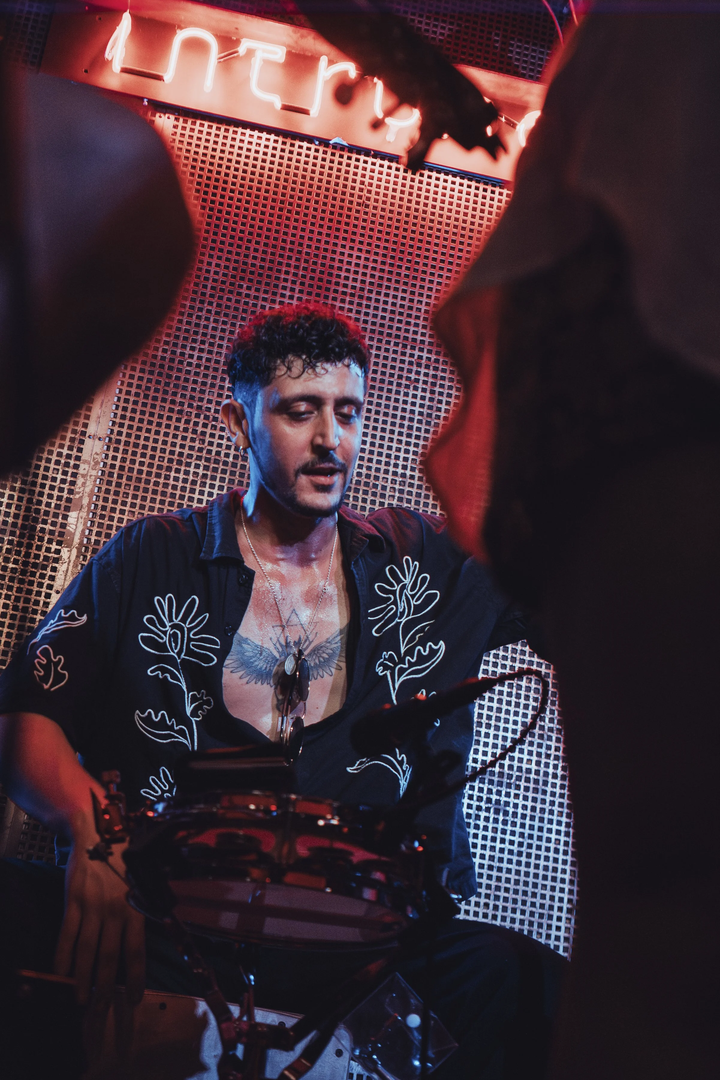 Un hombre tocando un tambor en un escenario con luces de neón y fondo de rejilla, con el cartel 'Library' parcialmente visible.