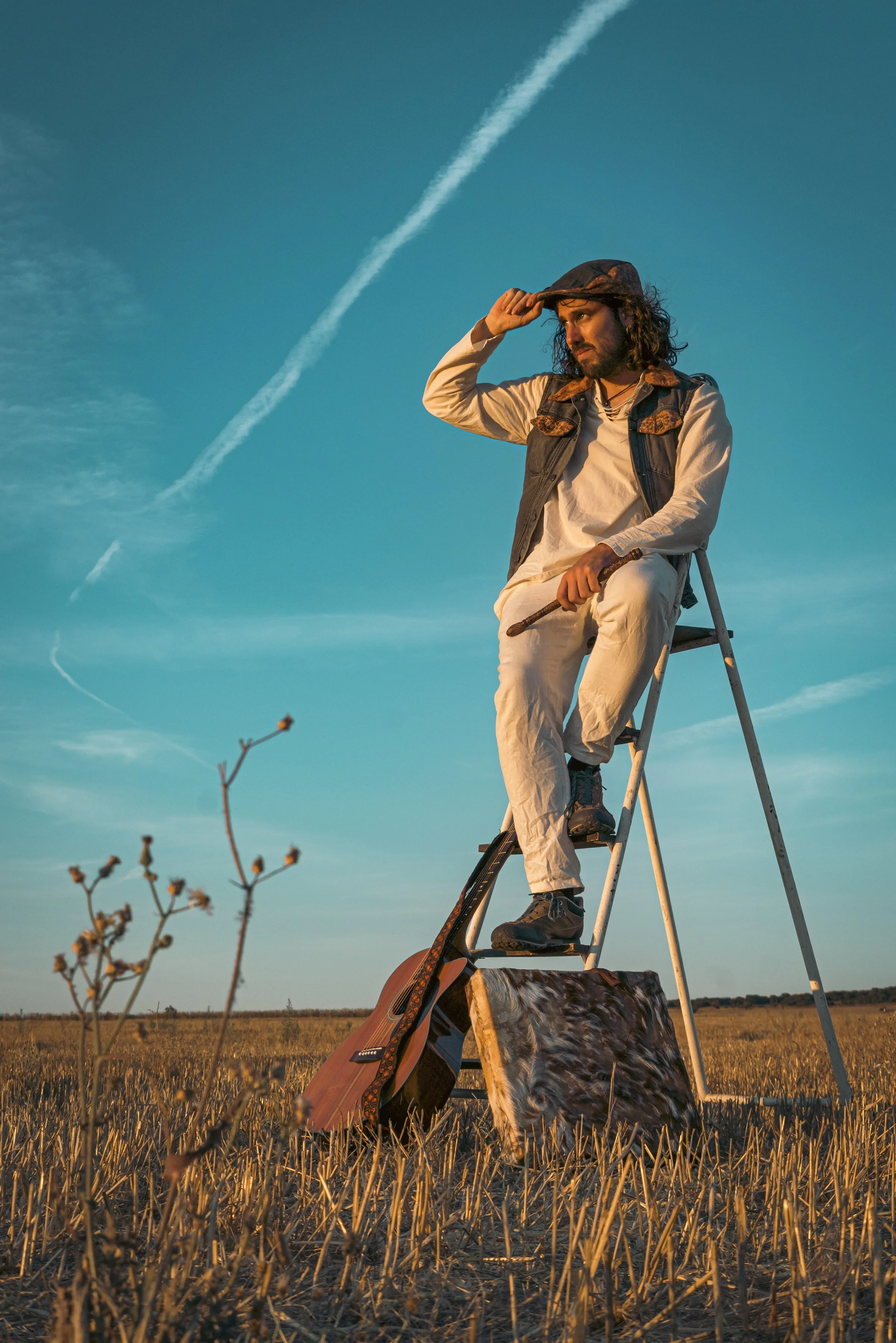 Hombre sentado en una escalera en un campo, con guitarra apoyada en la tierra, saludando y mirando hacia el horizonte al atardecer, con cielo despejado y contrails.
