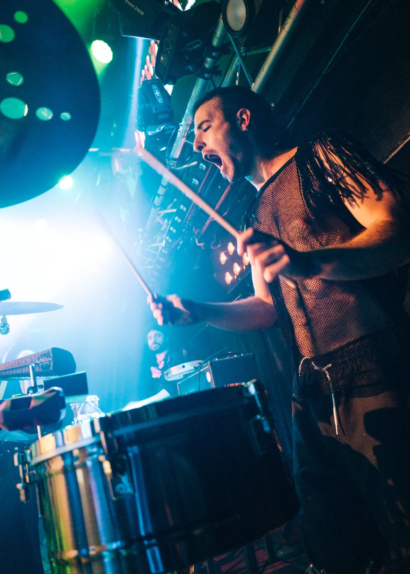 Una persona tocando los tambores en un escenario con iluminación de colores, vestida con ropa de malla y con expresión enérgica.