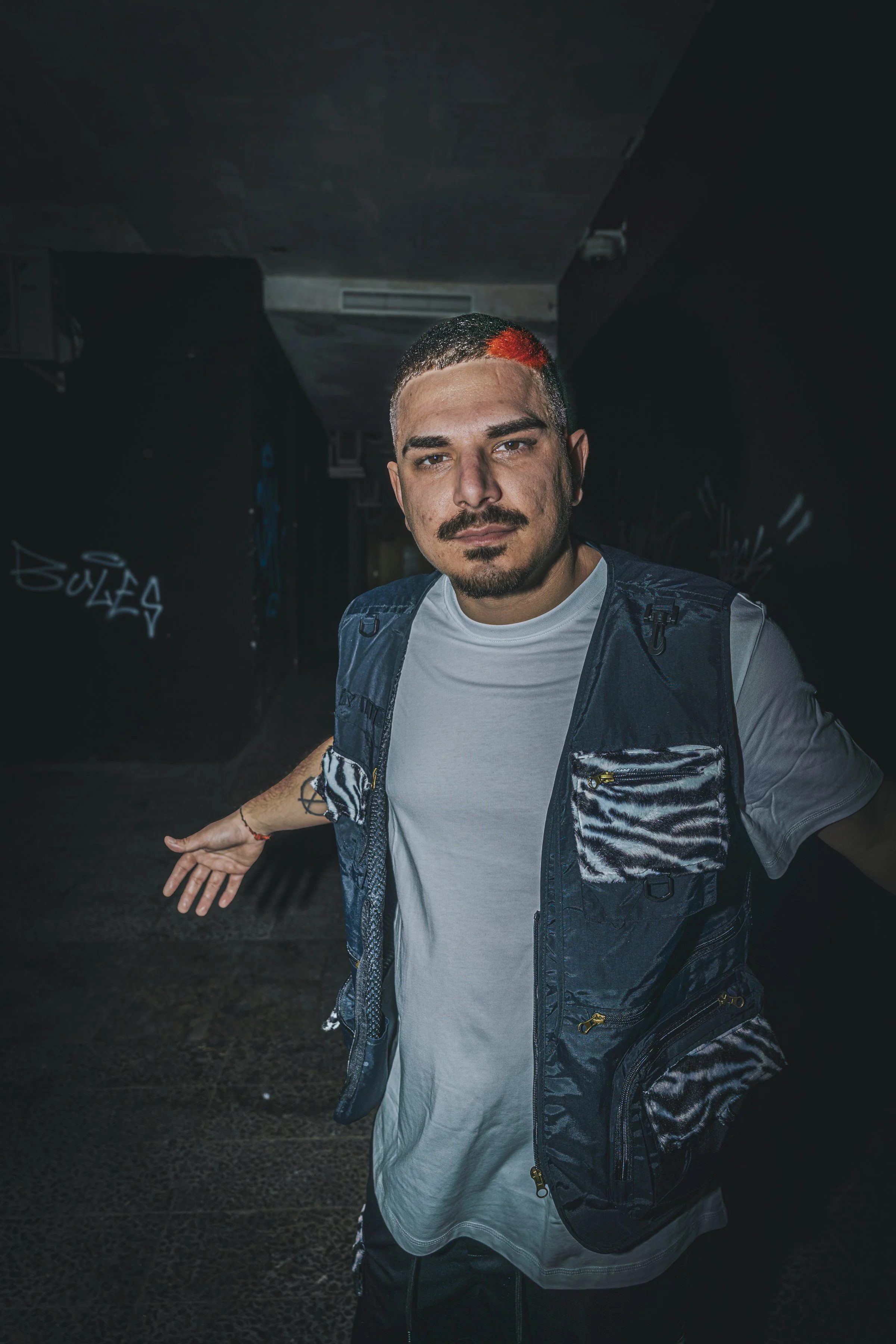 Joven con barba y cabello corto, con un toque de color rojo en el lado, vistiendo camiseta blanca y chaleco con bolsillos de felpa, posando en un espacio oscuro con graffiti en las paredes.
