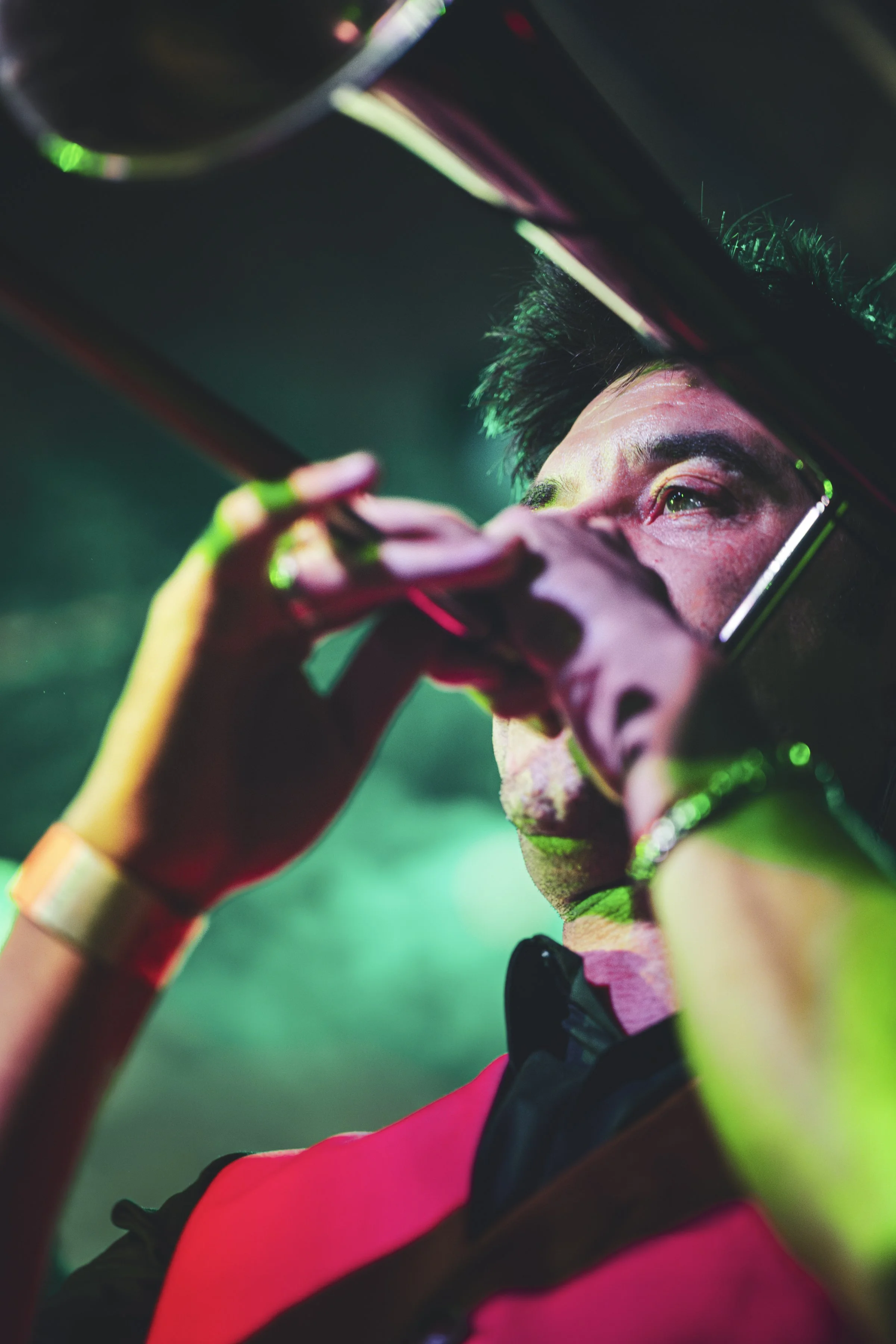 Joven con rostro emocionado, saludando y visto desde abajo, usando lentes oscuros y ropa deportiva, con fondo de luces verdes y rosas.