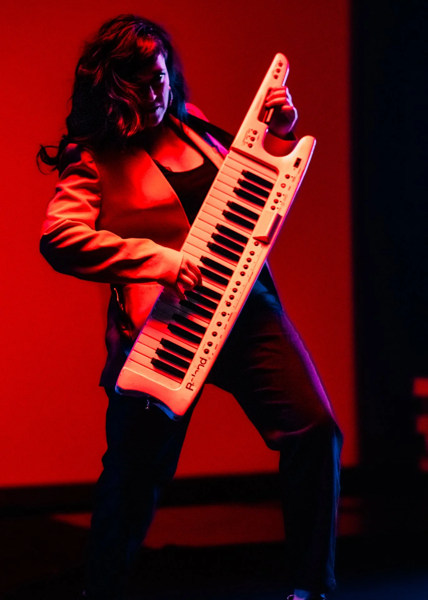 Mujer con cabello oscuro y ropa negra y roja, tocando un teclado musical eléctrico en un escenario con iluminación roja y oscura.
