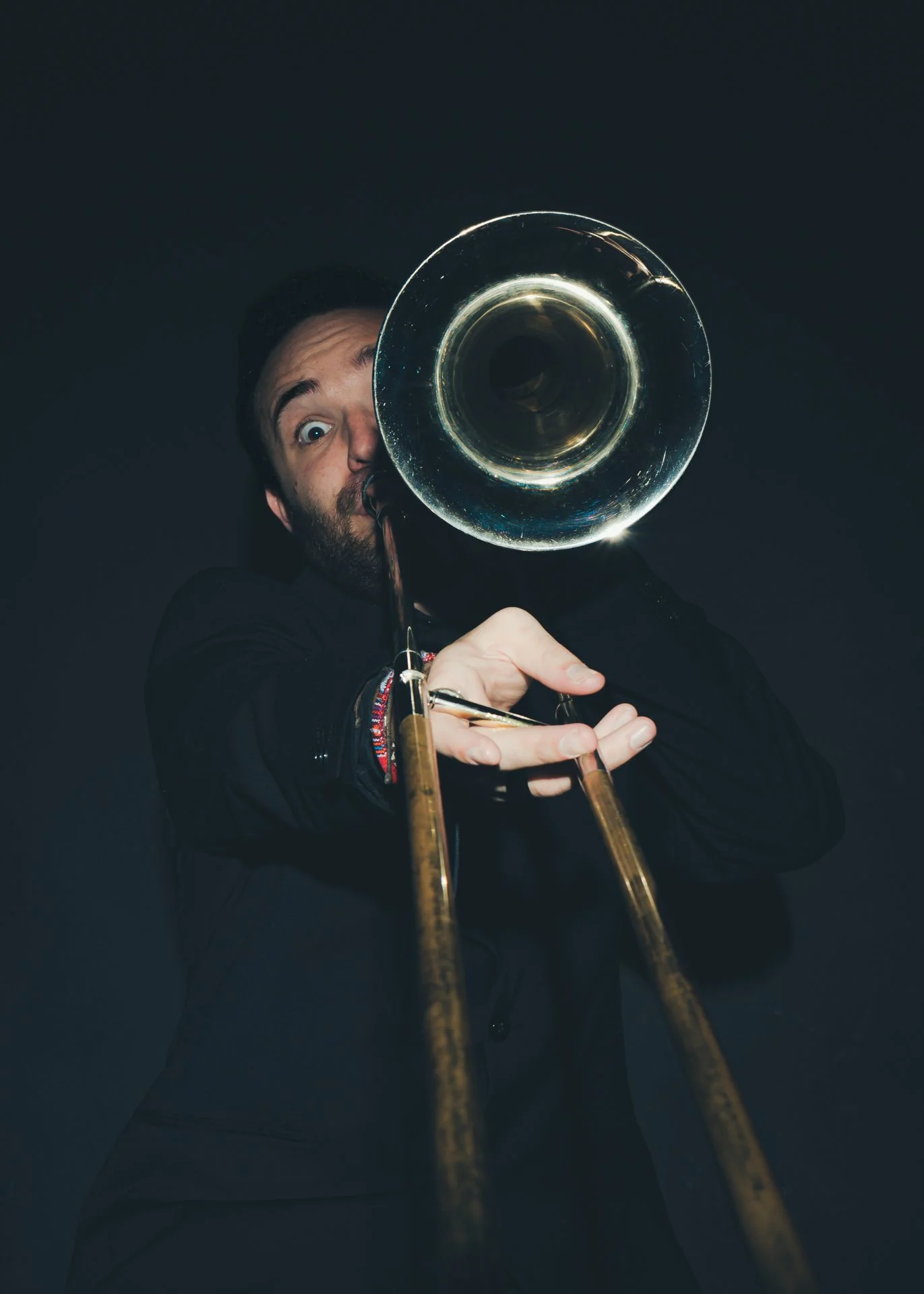 Un hombre tocando trombón, vestido de negro, con expresión divertida y fondo oscuro.