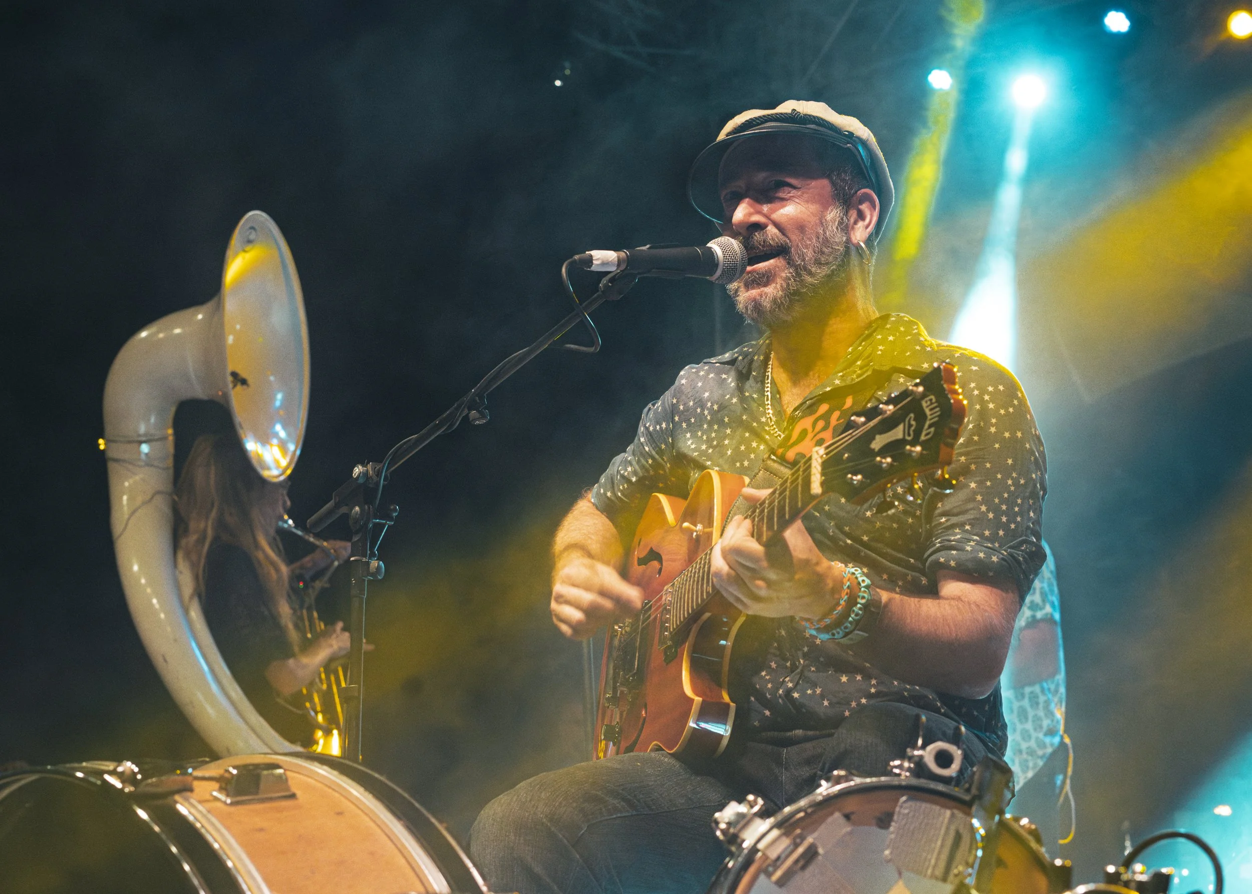Hombre tocando guitarra y cantando en un escenario con luces de colores, con un micrófono y un megáfono delante.