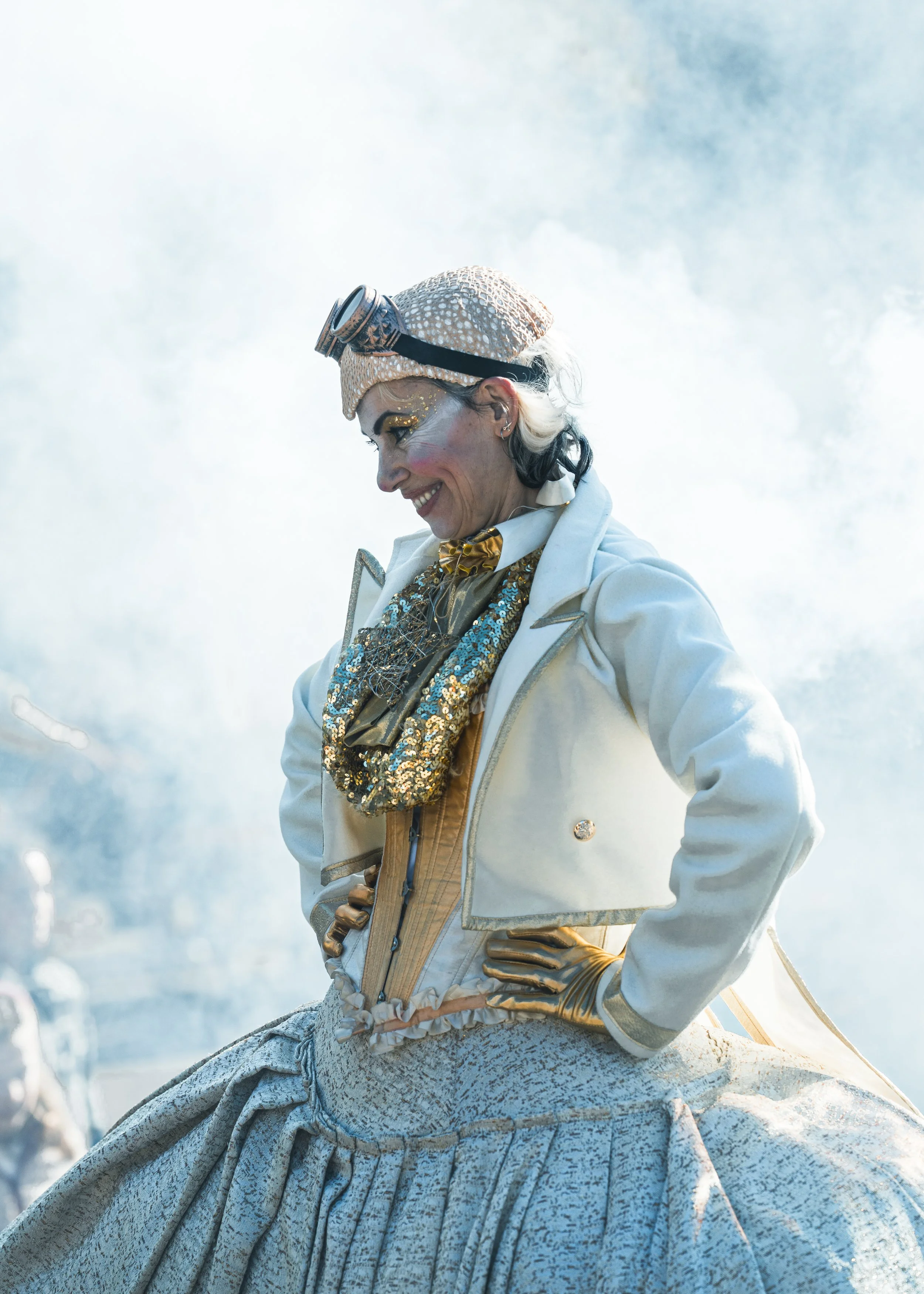 Una mujer con vestimenta colorida, con un sombrero y accesorios brillantes, sonriendo y posando en un entorno con humo de fondo.