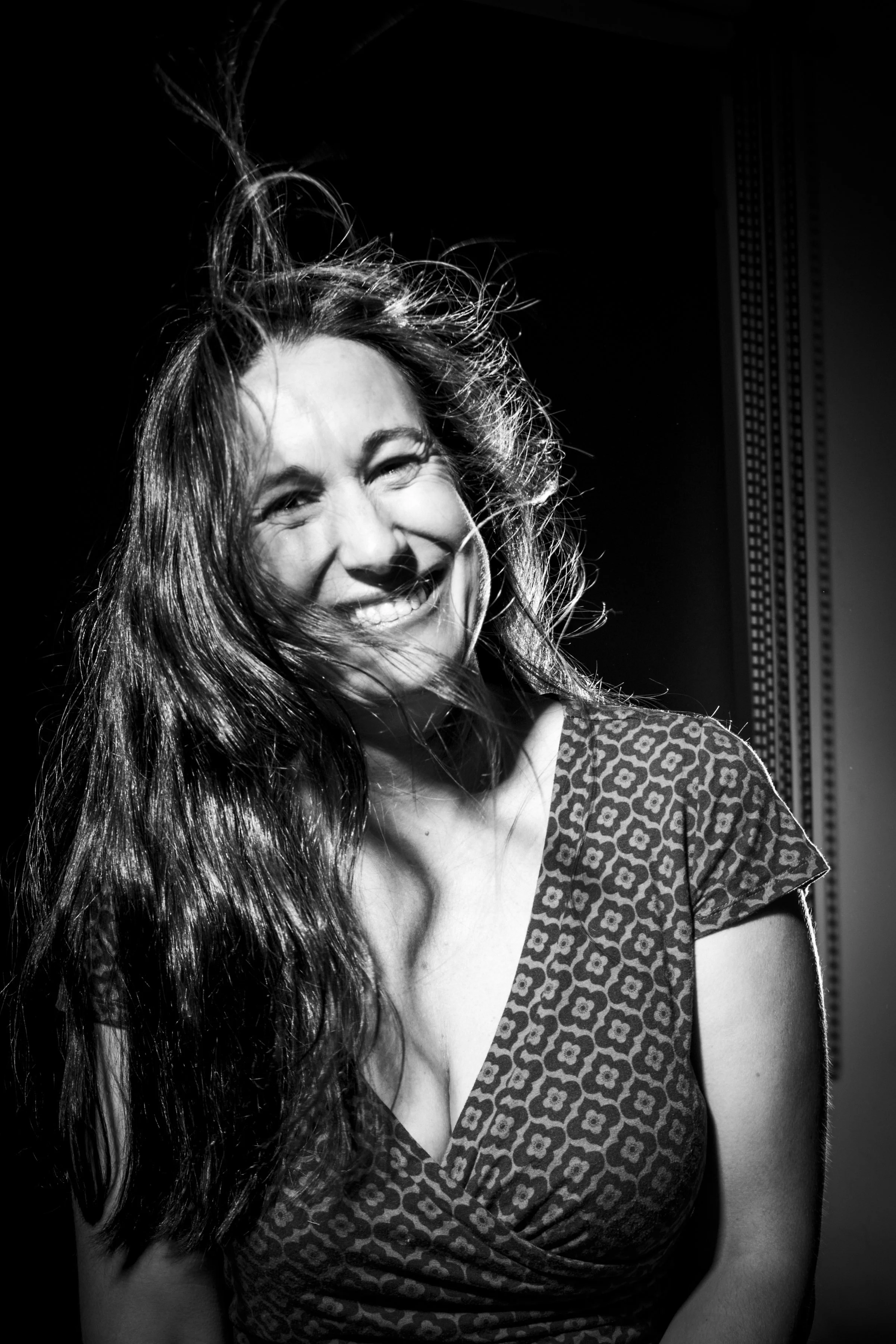 Retrato en blanco y negro de una mujer sonriendo, con cabello largo y ondulado, usando un vestido con estampado de flores, en un fondo oscuro con luz que ilumina su rostro y cabello.