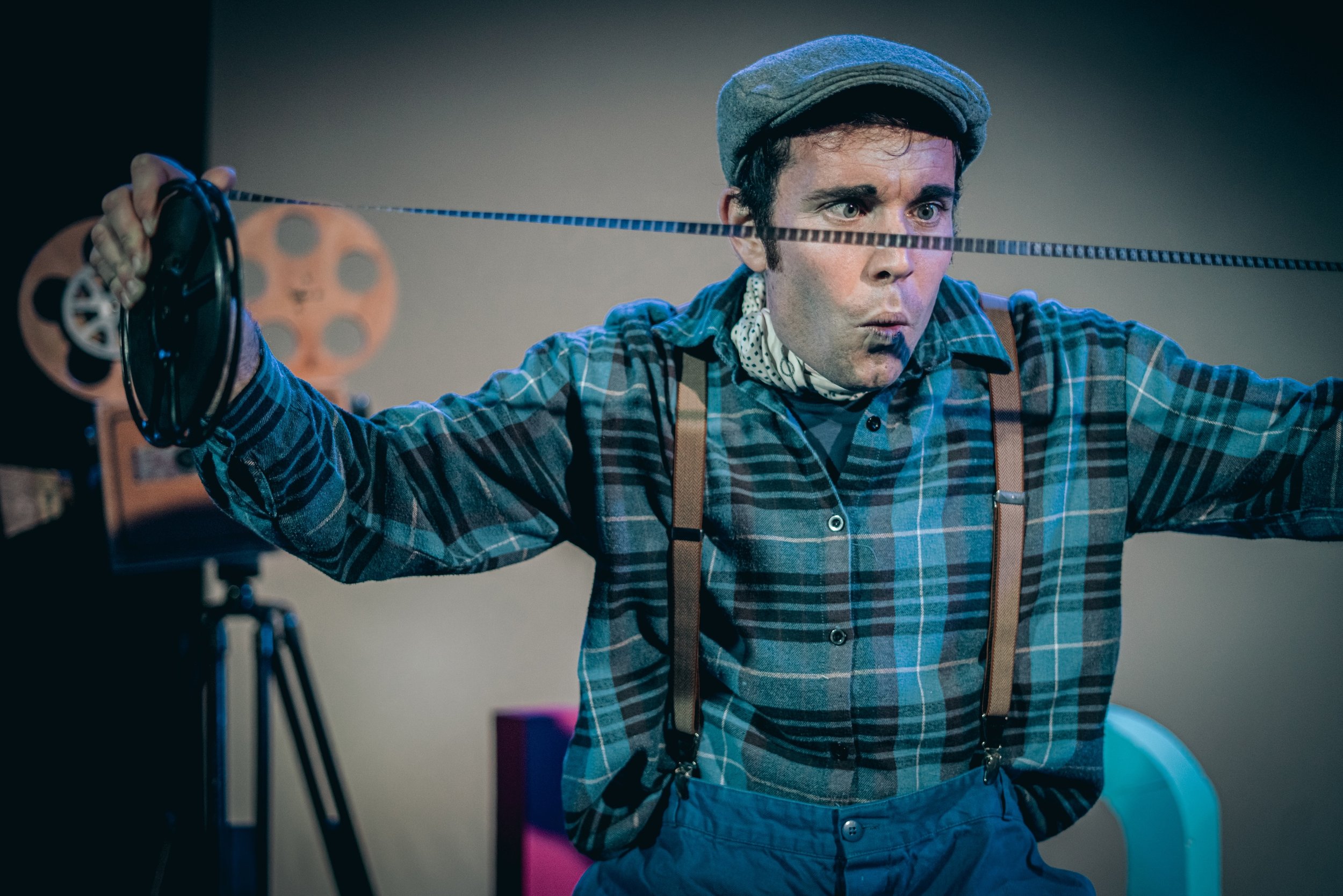 Un hombre actuando como cineasta, ajustando una cinta de film delante de un proyector, usando ropa de estilo vintage y un gorro, en un ambiente de teatro o estudio de cine.