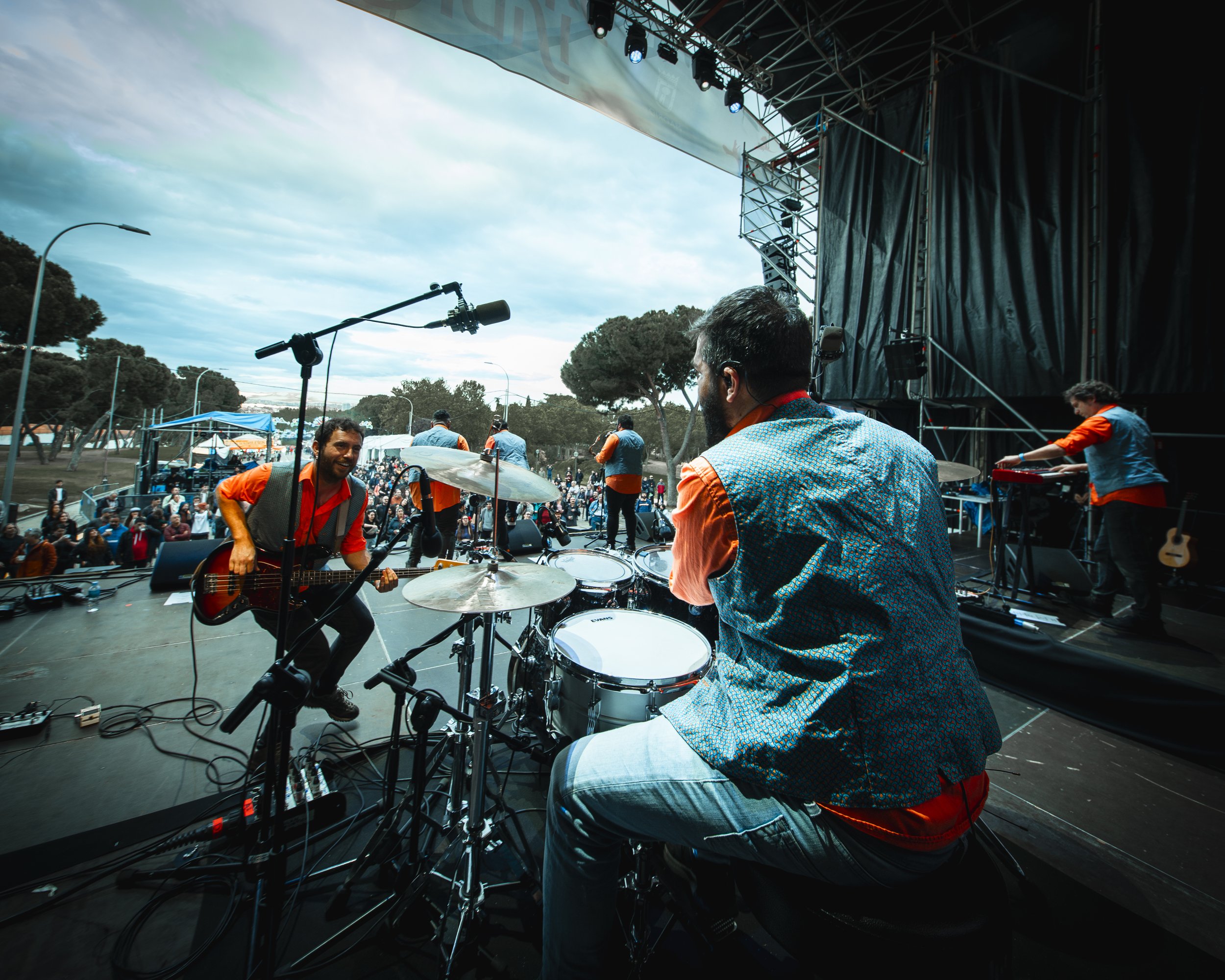 Banda tocando en un escenario al aire libre durante un concierto, con público asistente y árboles en el fondo.