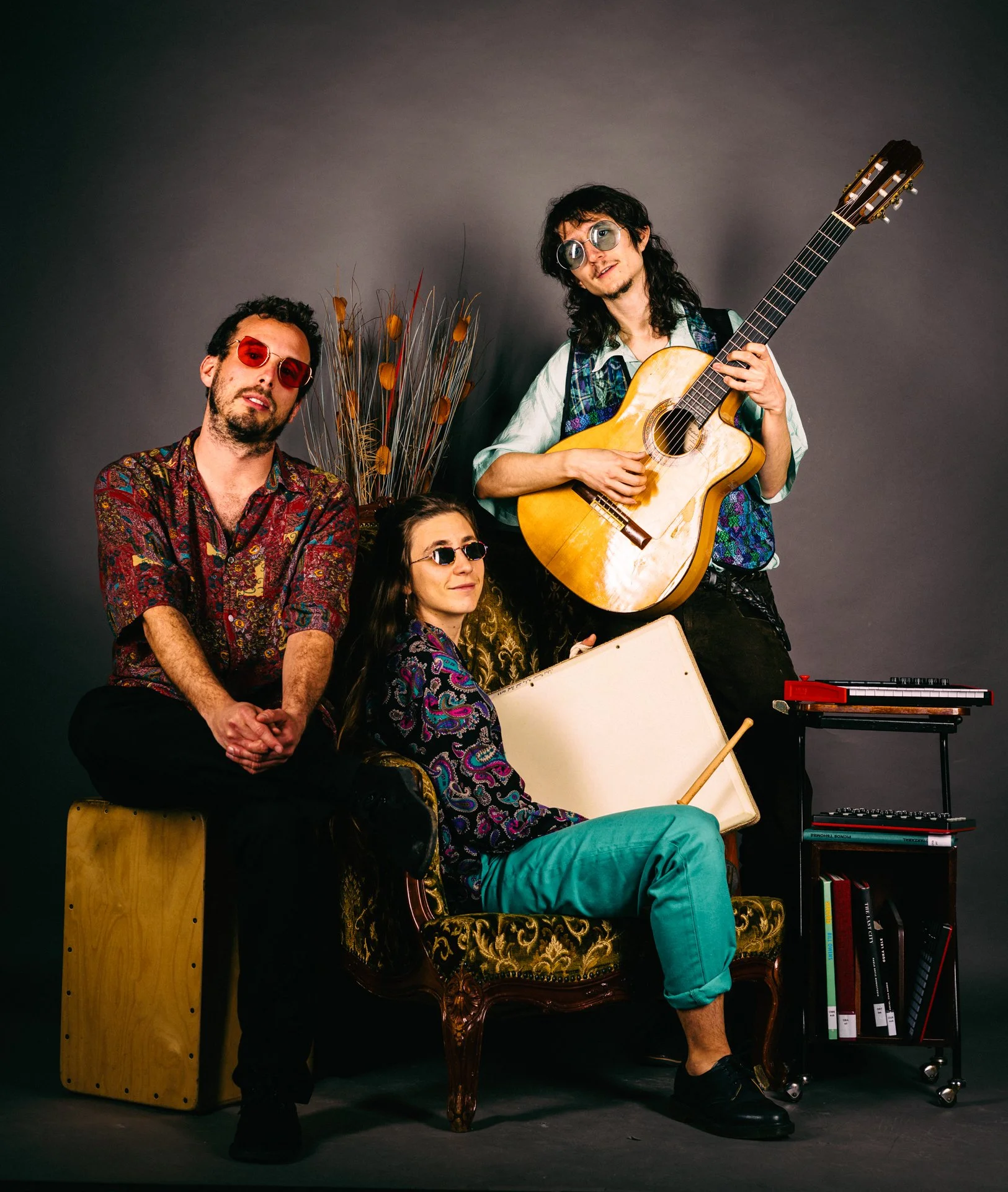 Cuatro personas con gafas de sol y ropa colorida posando con instrumentos musicales en un estudio con fondo gris oscuro.