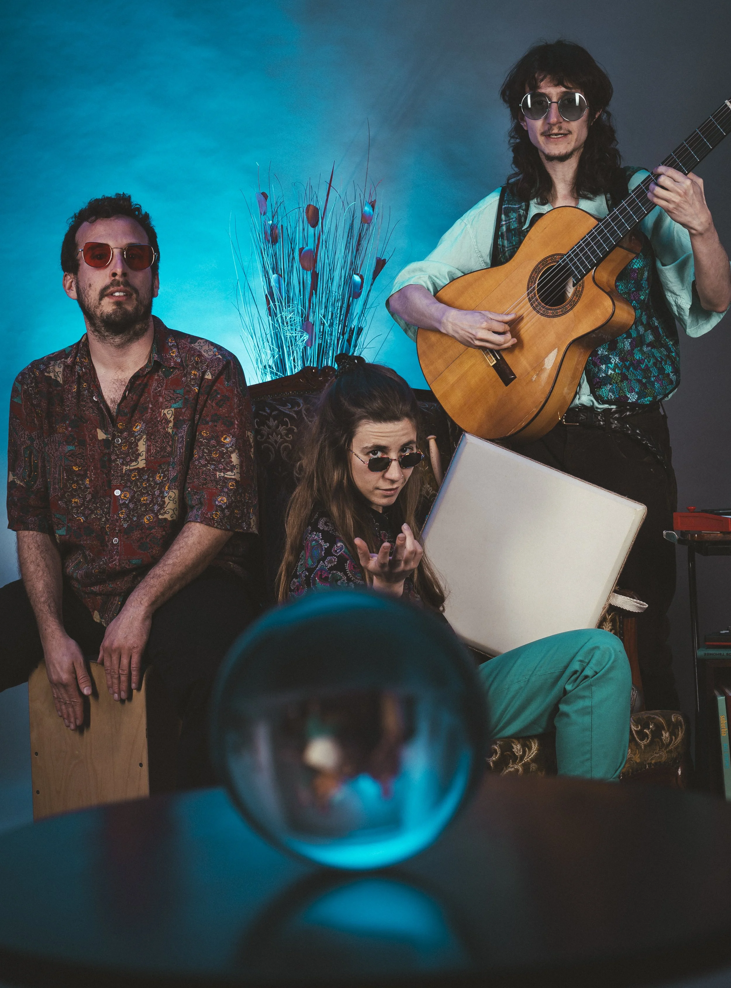 Tres personas en un ambiente con iluminación azul, una mujer sentado en un sillón con gafas de sol, otra mujer con gafas en el suelo gesticulando, un hombre con gafas de sol tocando la guitarra, y un objeto reflectante en primer plano.