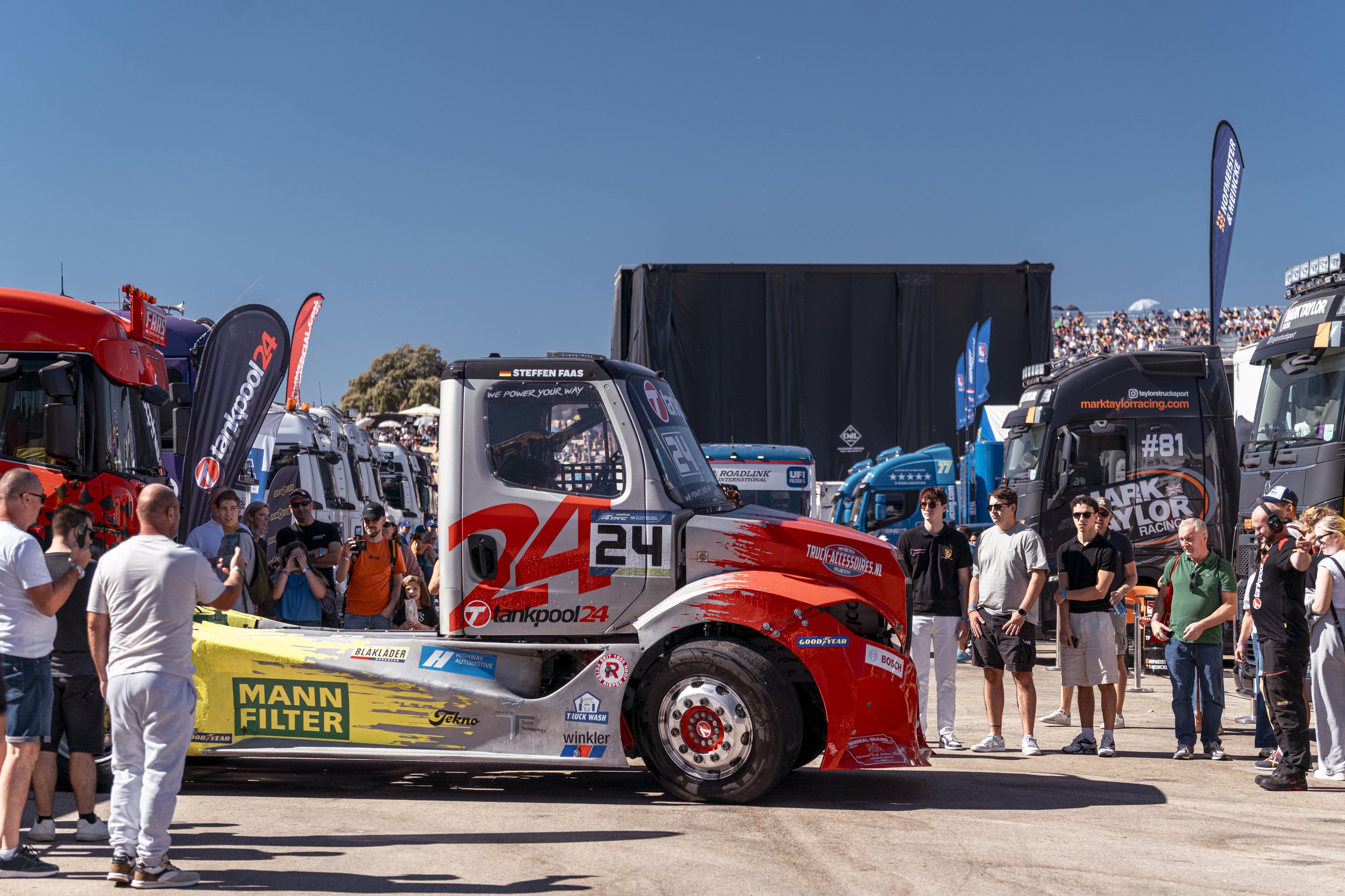 Gran camión de carreras en un evento, rodeado de espectadores y otros camiones en el paddock, durante un día soleado.